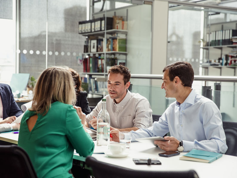 Four people sit around a table in a modern office, engaged in discussion. Papers, notebooks, and a water bottle are on the table, and shelves with books and files are visible in the background.