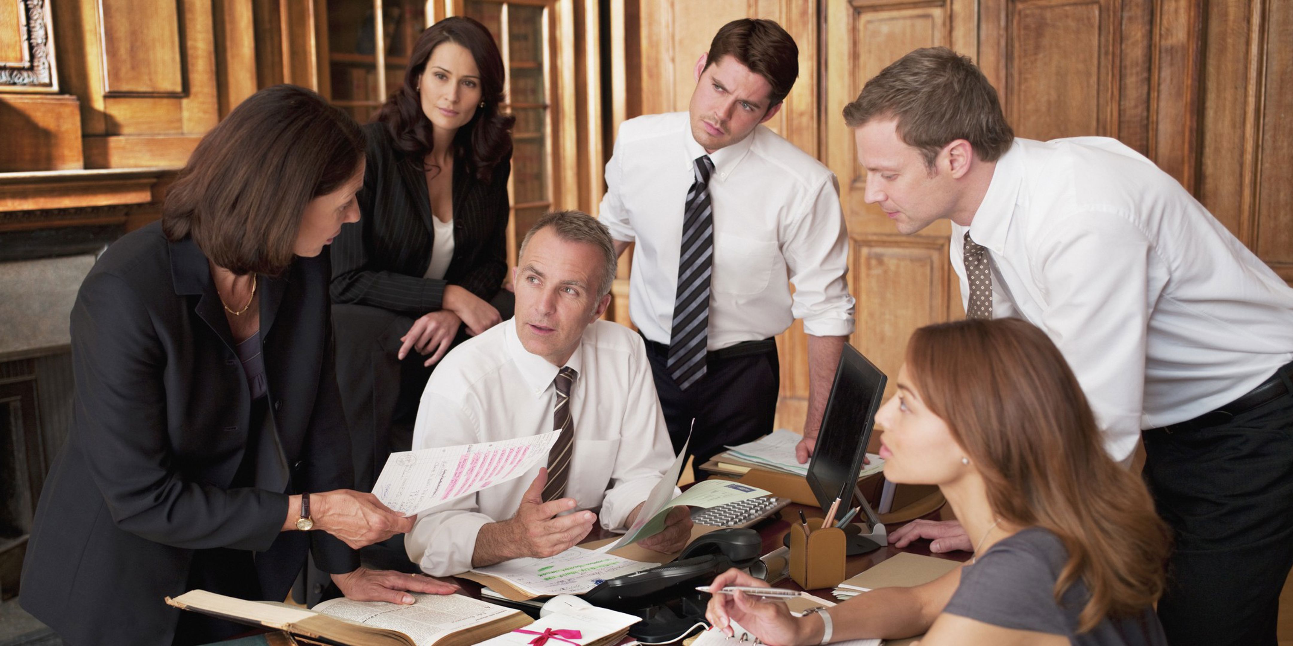 Five people in business attire have a discussion around a cluttered office desk with papers, files, a computer, and a phone, appearing focused and engaged in conversation. The setting is a wood-paneled office.