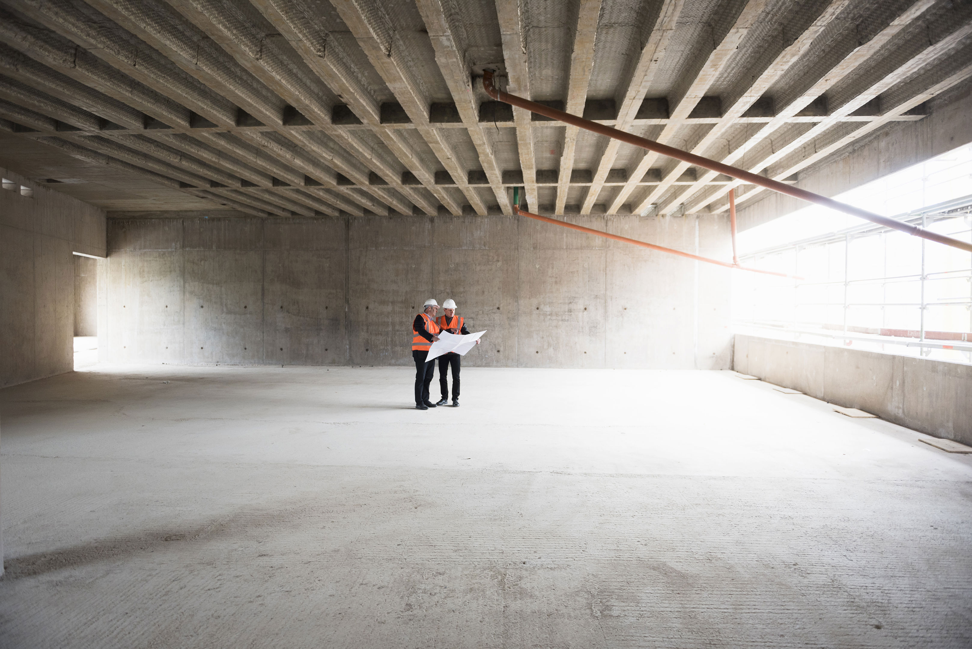 Two Men With Plan Wearing Safety Vests Talking In Building Under Construction