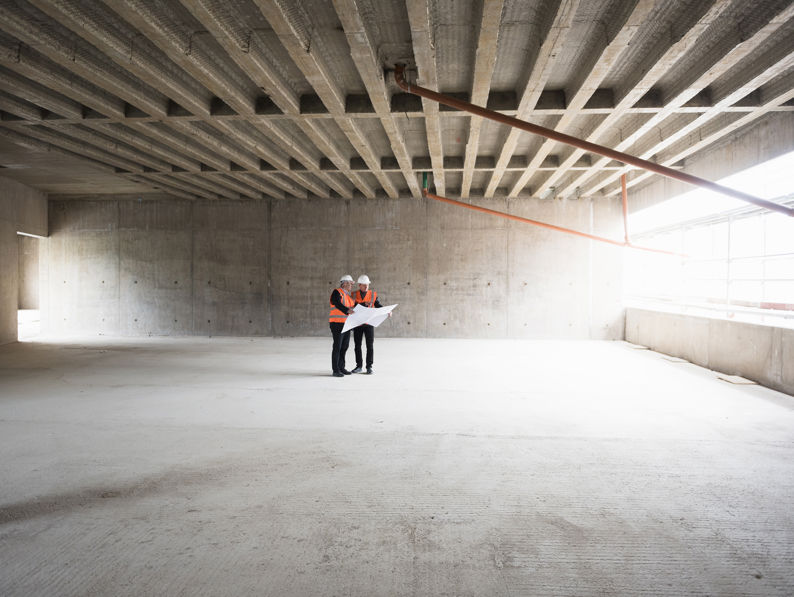 Two Men With Plan Wearing Safety Vests Talking In Building Under Construction