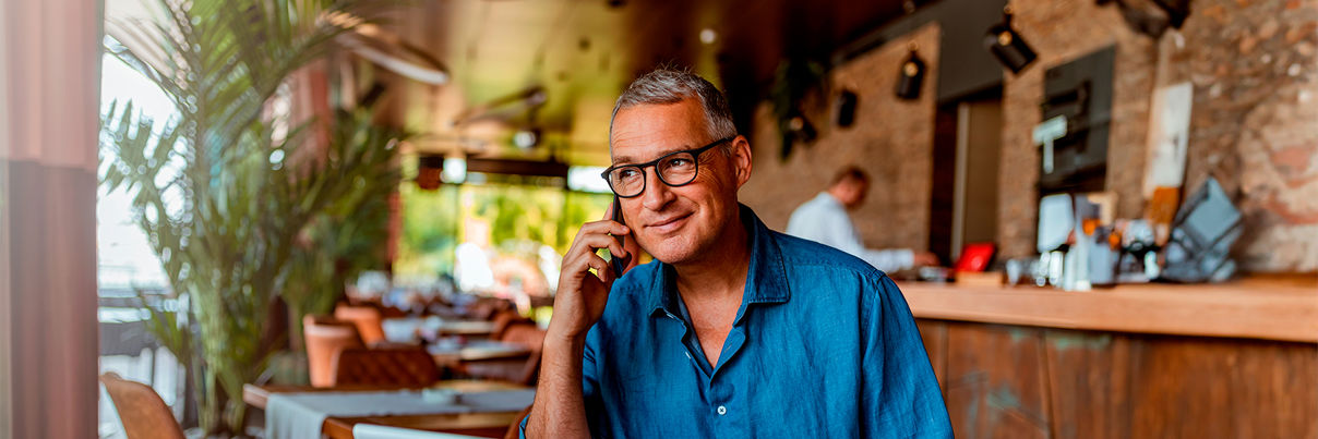 A man in a blue shirt is sitting at a cafe table with a laptop and a drink. He is smiling and talking on the phone. The cafe has wooden decor, shelves with bottles, and a row of tables. Lush green plants are in the background.