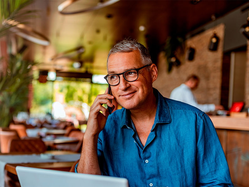 A man in a blue shirt is sitting at a cafe table with a laptop and a drink. He is smiling and talking on the phone. The cafe has wooden decor, shelves with bottles, and a row of tables. Lush green plants are in the background.