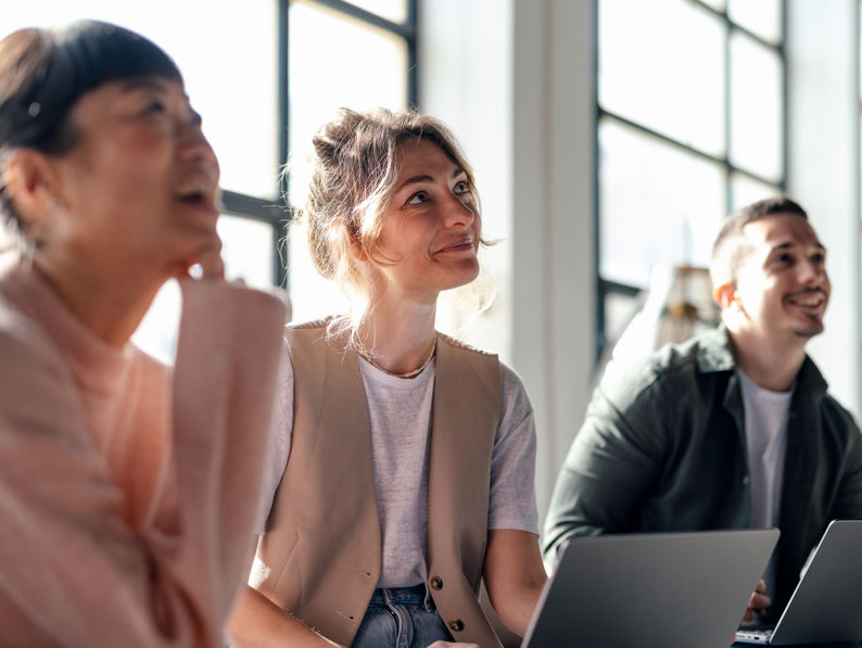 Three people sit indoors, smiling and looking in the same direction. They are casually dressed and have laptops open in front of them. The room is bright with large windows in the background.