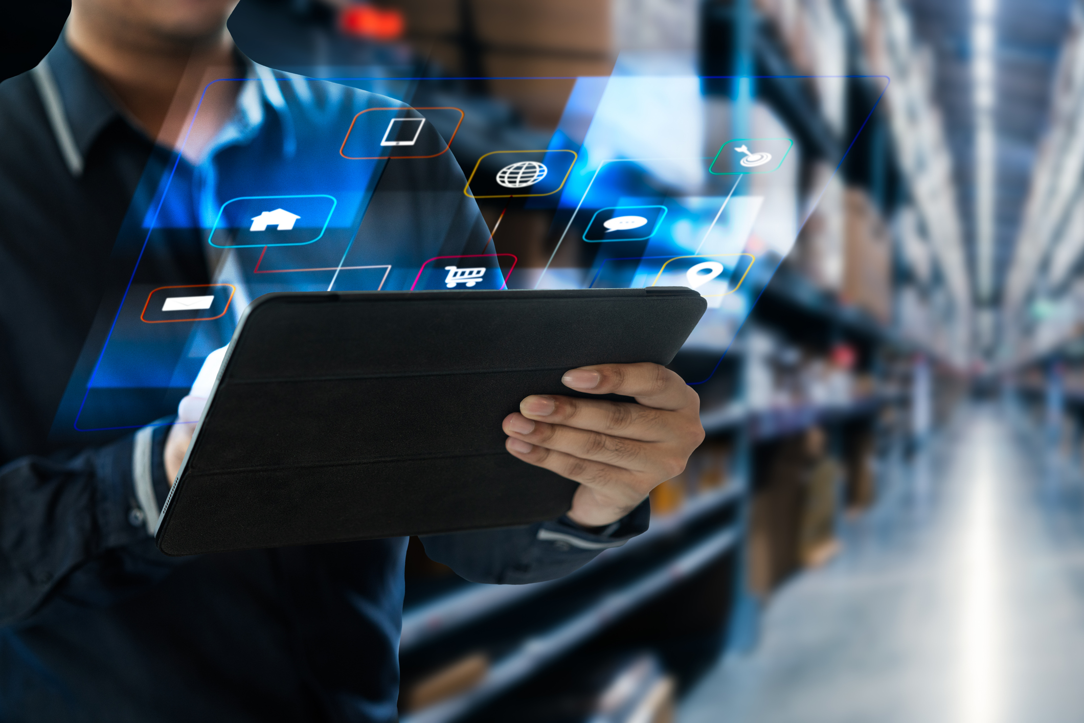 A person uses a tablet with digital icons projected above it, standing in a warehouse aisle filled with shelves and cardboard boxes. The scene suggests technology integration in logistics or inventory management.