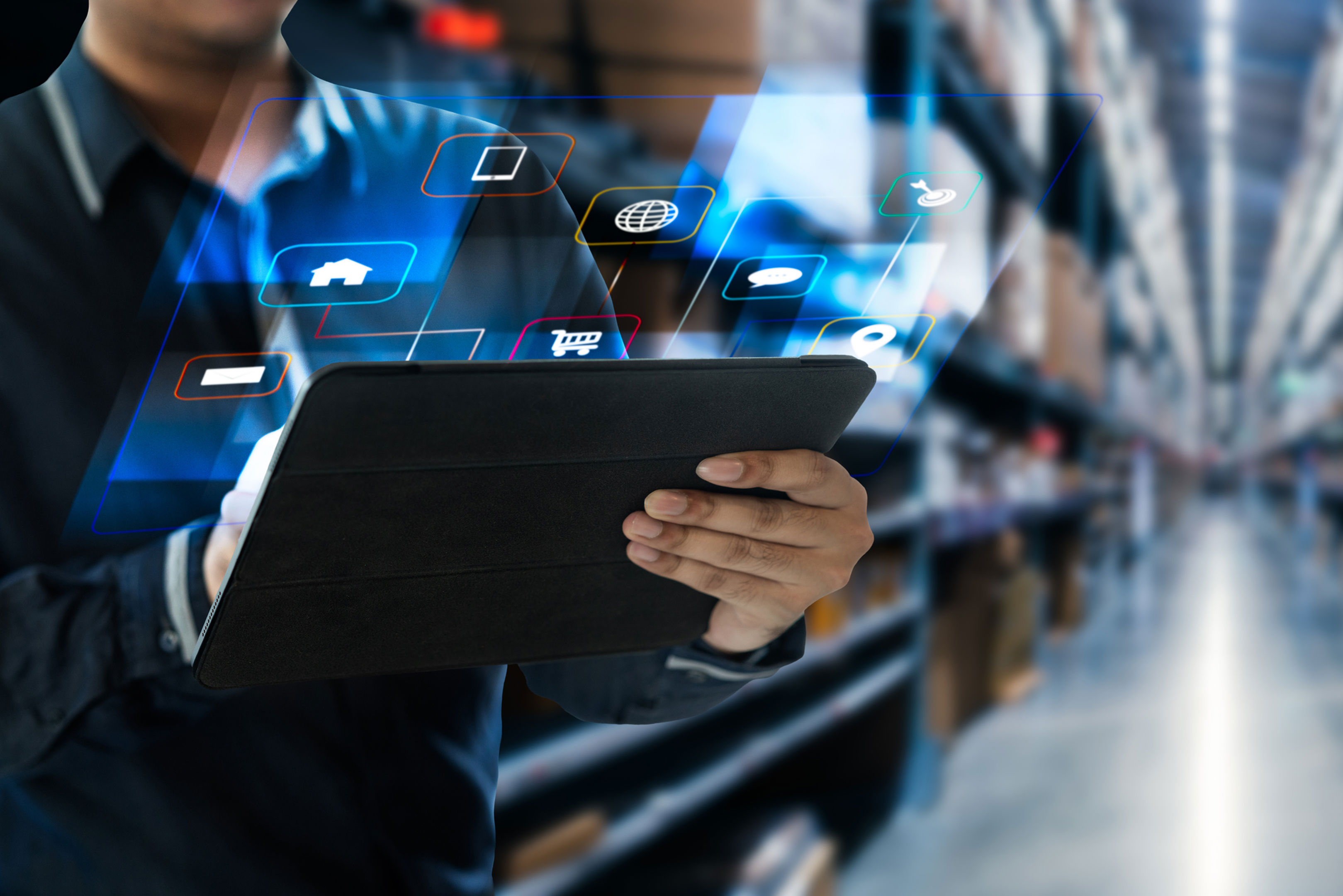 A person uses a tablet with digital icons projected above it, standing in a warehouse aisle filled with shelves and cardboard boxes. The scene suggests technology integration in logistics or inventory management.