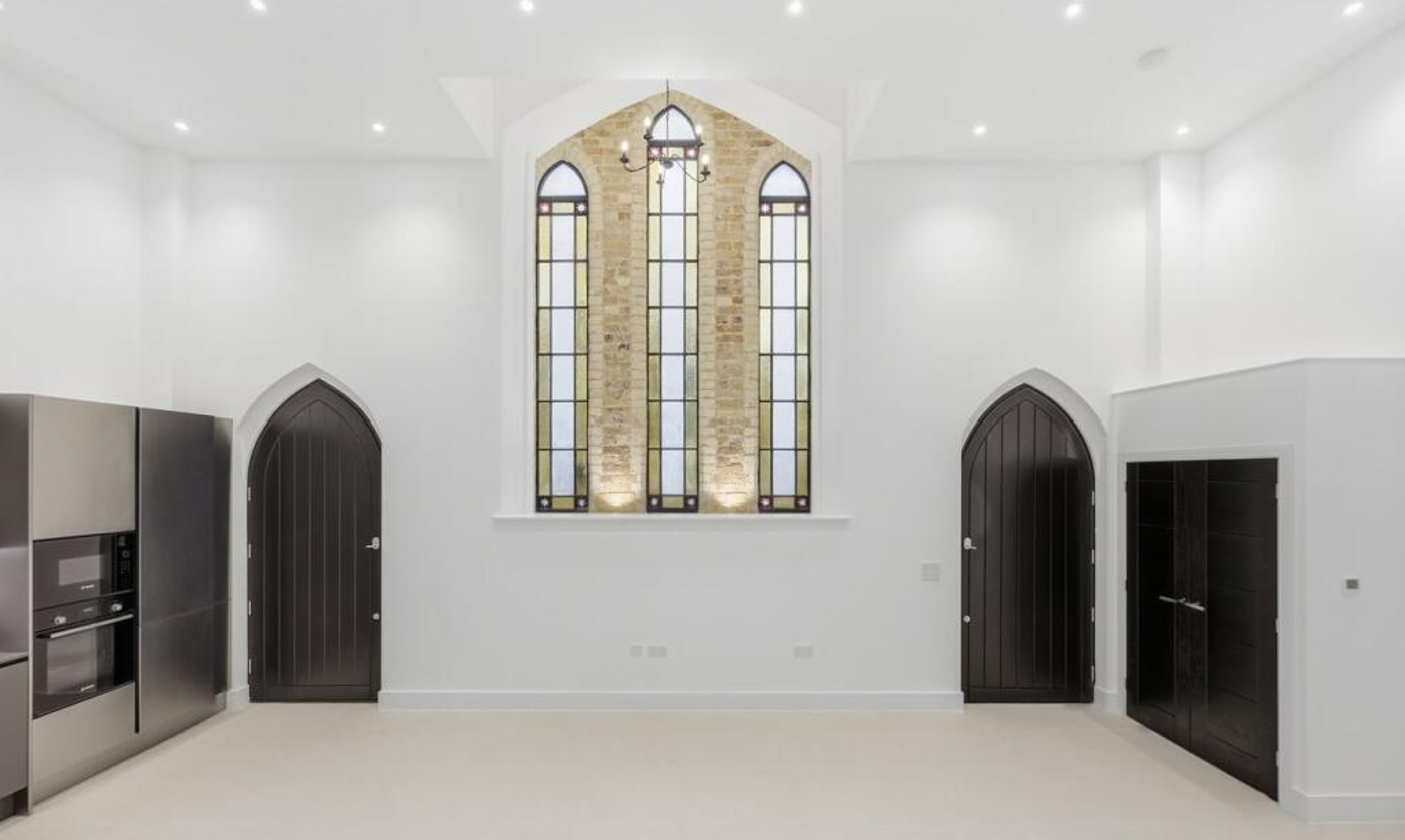 A modern kitchen in a converted church features tall arched windows with stained glass, black arched doors, white walls, and built-in stainless steel appliances. Minimalist and bright with high ceilings.