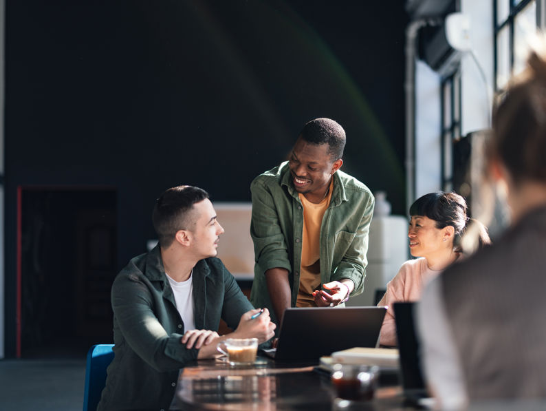Four people are gathered around a table in a bright, modern office. Three are engaged in conversation, smiling, with laptops and coffee cups in front of them. One person is blurred in the foreground, facing away.