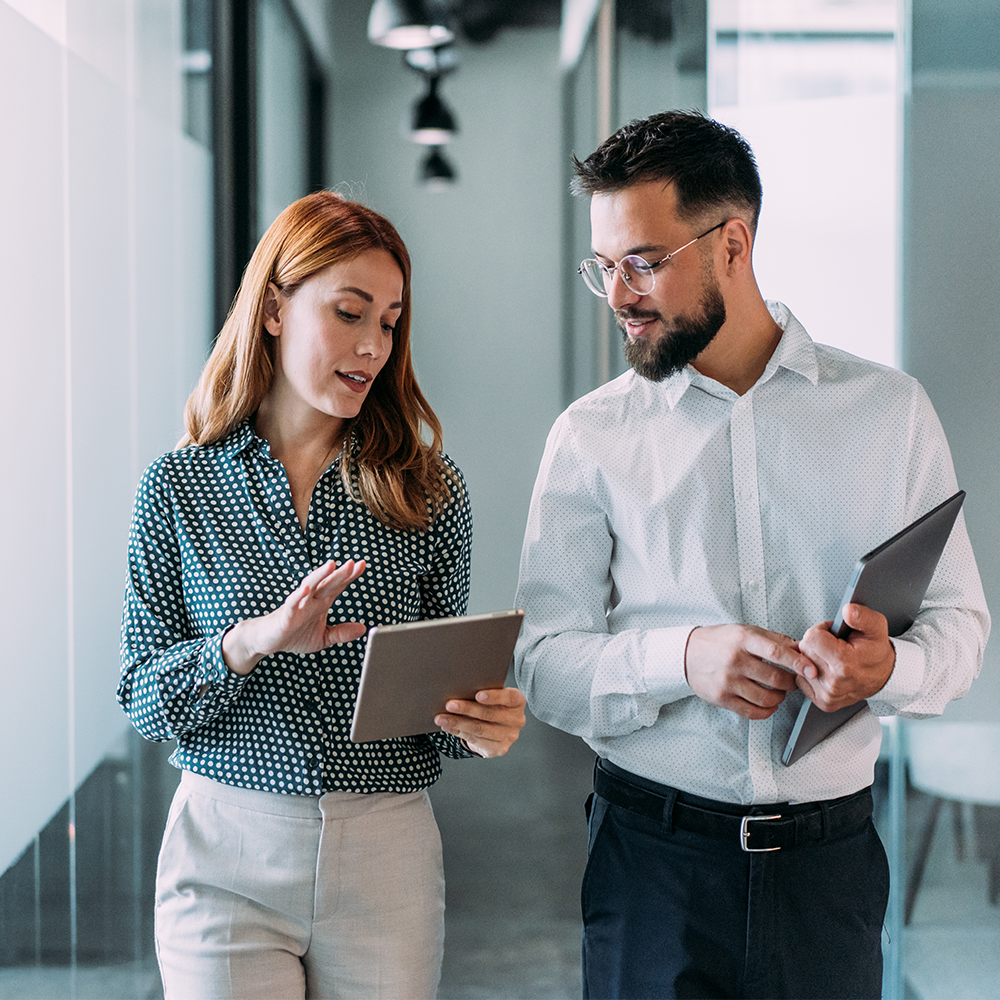 A man and a woman in business attire walking through an office while discussing information on a tablet.