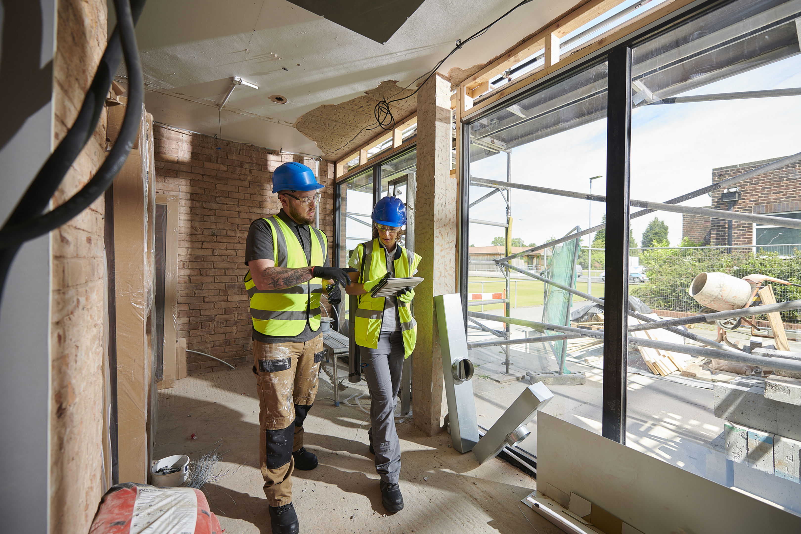 Two construction workers in safety vests and blue helmets discuss plans while standing inside a partially built structure with large glass windows and building materials visible.