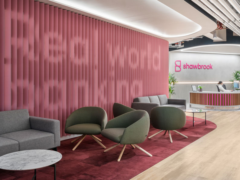 Modern office lobby with pink accent wall displaying "Real world banking," gray and green lounge chairs, a round marble table, and a reception desk with the Shawbrook logo in the background.
