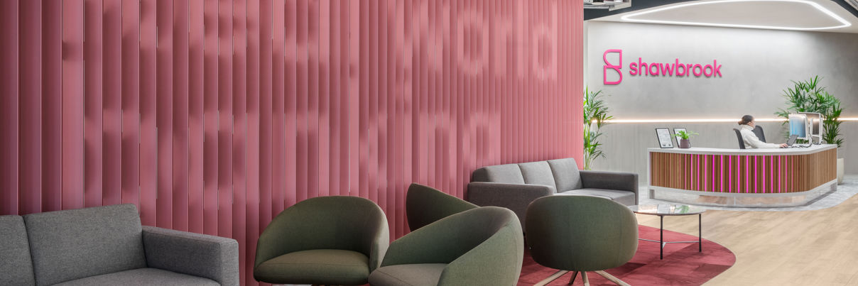 Modern office lobby with pink accent wall displaying "Real world banking," gray and green lounge chairs, a round marble table, and a reception desk with the Shawbrook logo in the background.