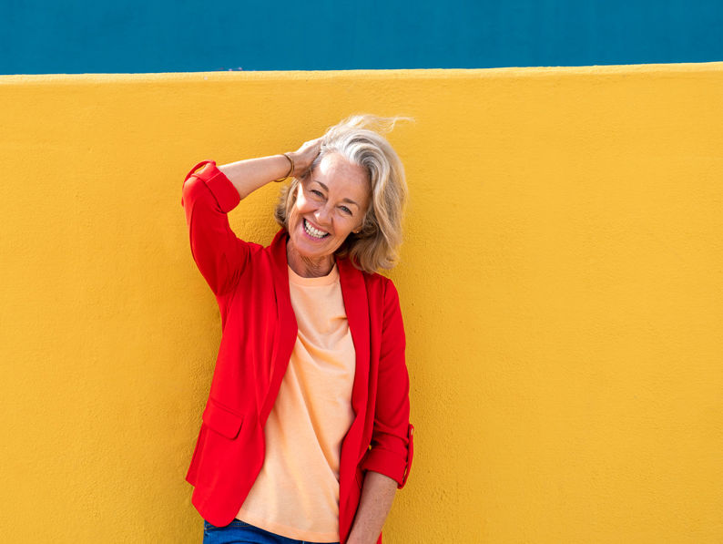 A smiling older woman with blonde hair, wearing a red blazer and light orange shirt, stands against a bright yellow wall with a teal stripe above, holding one hand behind her head.