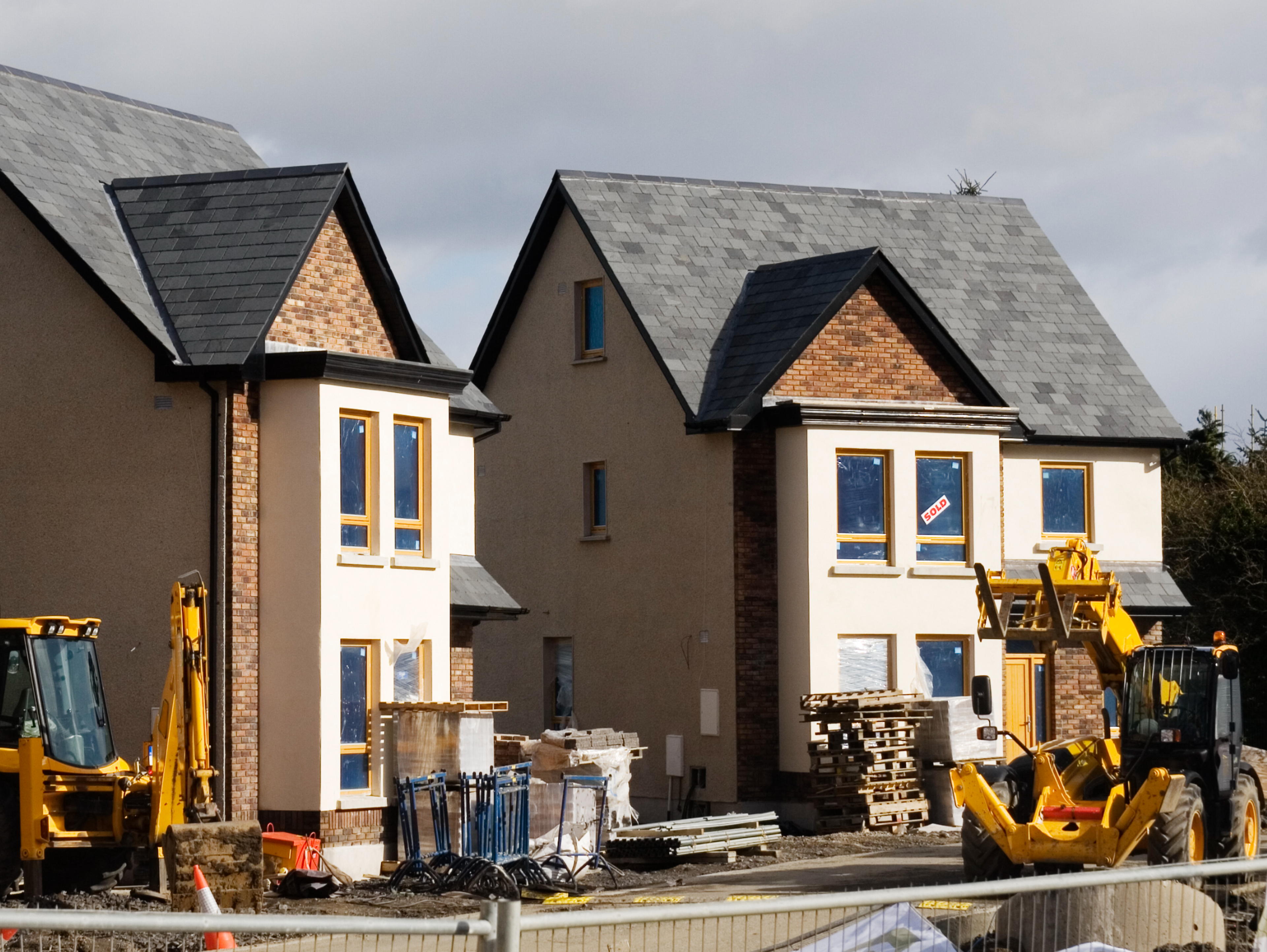 Two modern houses under construction with unfinished yards, surrounded by construction equipment, pallets, and building materials on a cloudy day.