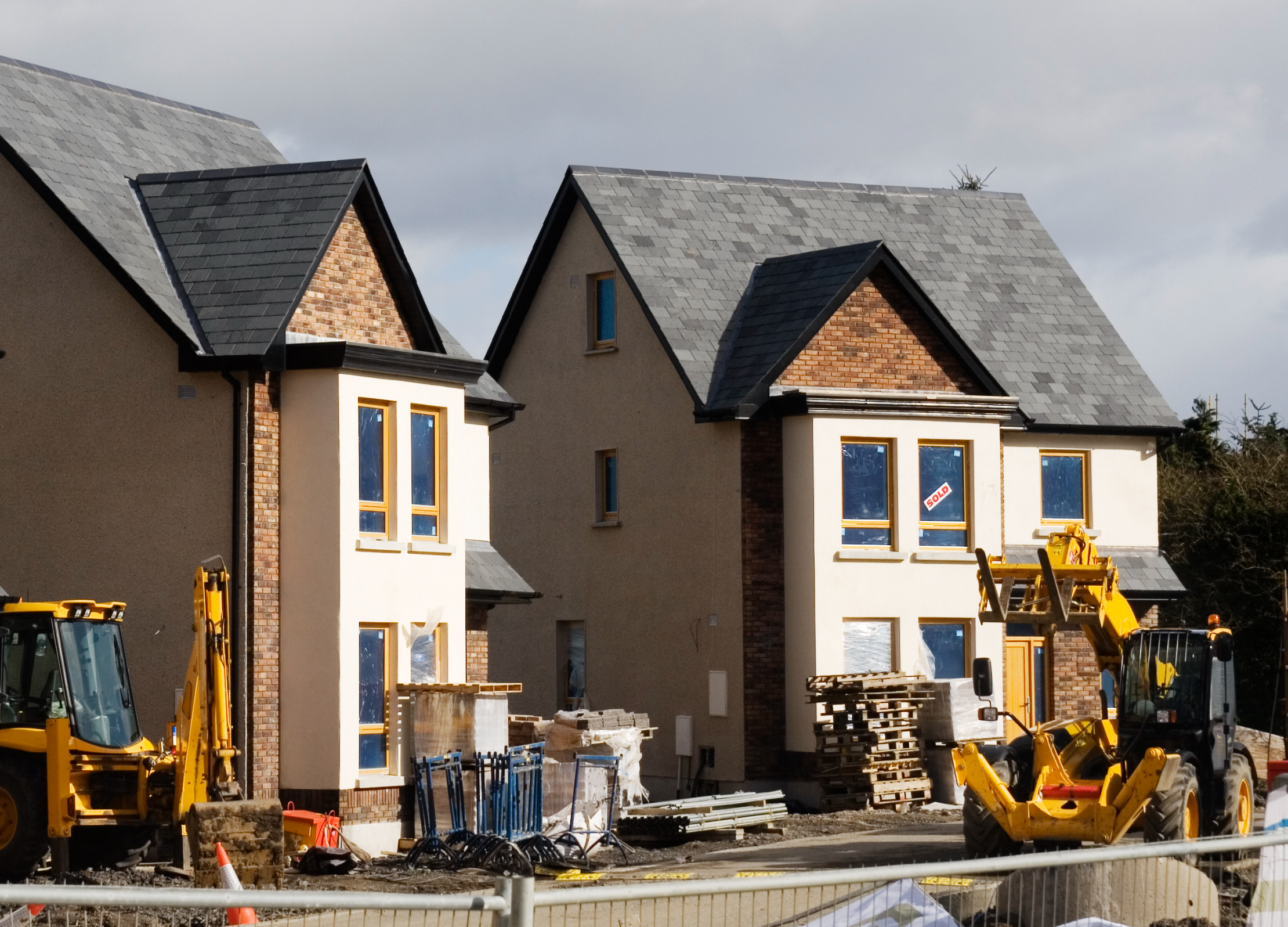 Two modern houses under construction with unfinished yards, surrounded by construction equipment, pallets, and building materials on a cloudy day.