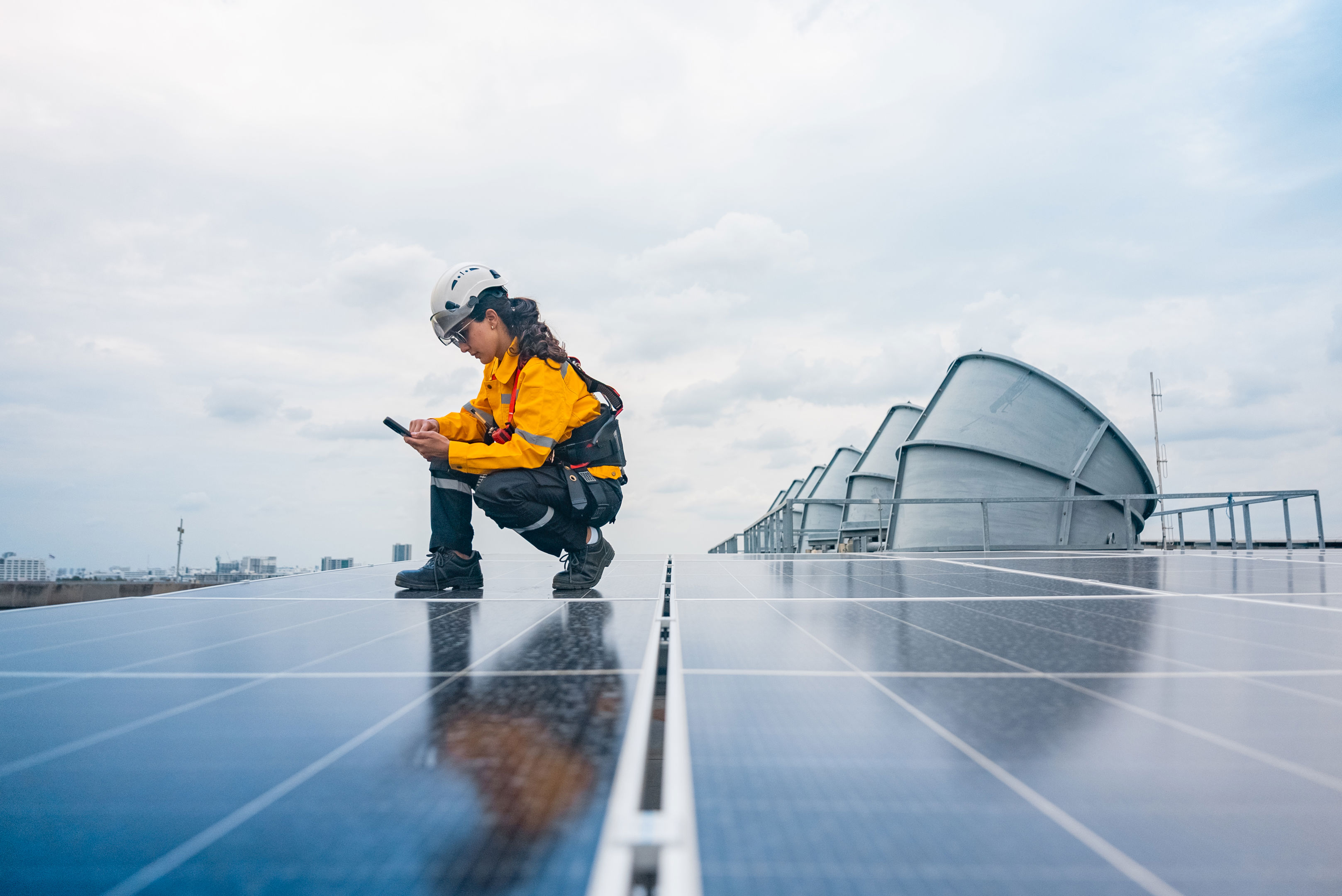 A person in safety gear crouches on solar panels, inspecting or using a device, with large ventilation units and a cloudy sky in the background.