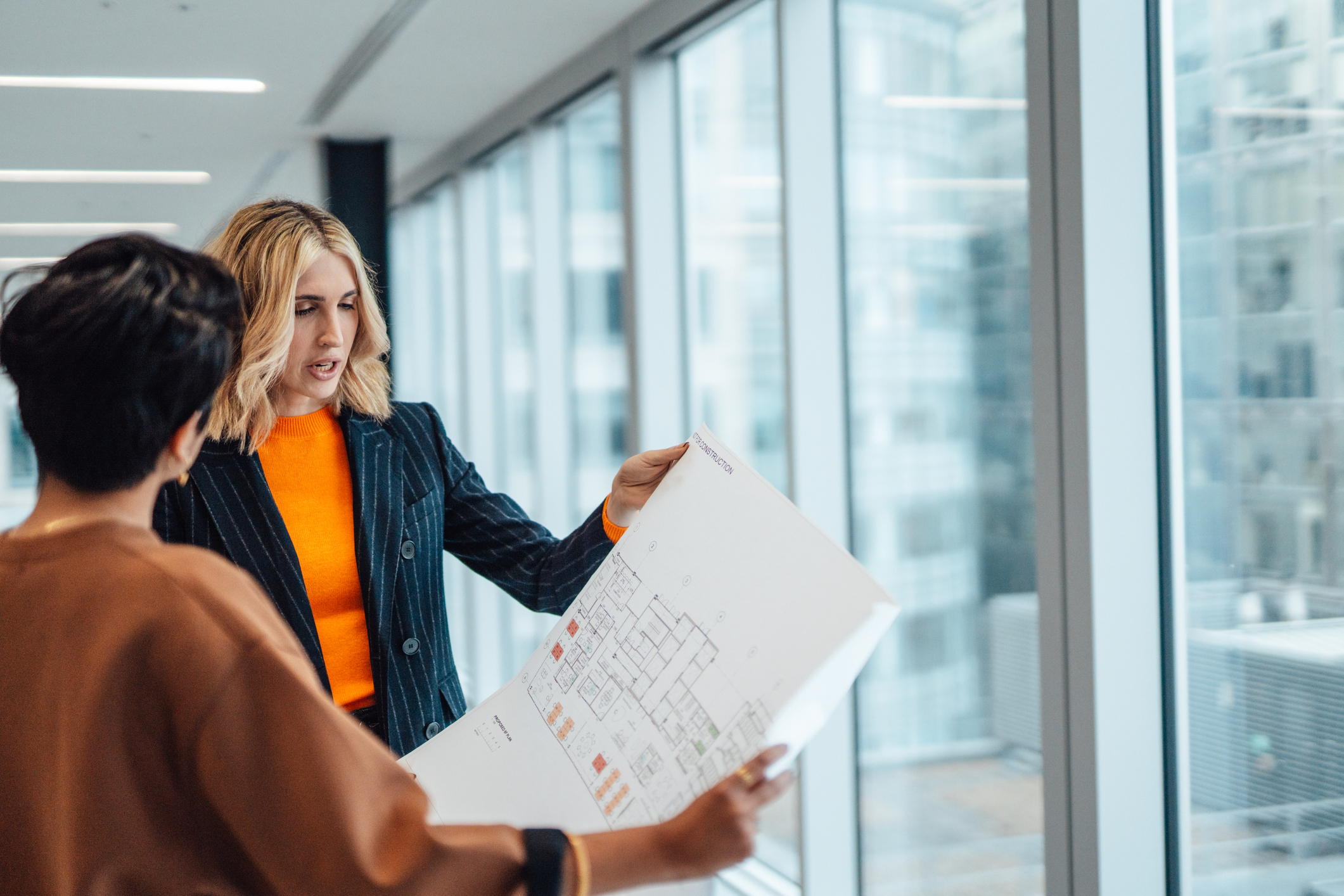 Two women stand by large office windows, discussing a printed architectural floor plan. One woman gestures and explains details, while the other listens and holds the plan.