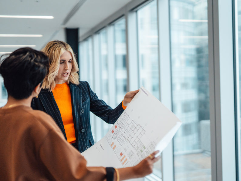 Two women stand by large office windows, discussing a printed architectural floor plan. One woman gestures and explains details, while the other listens and holds the plan.