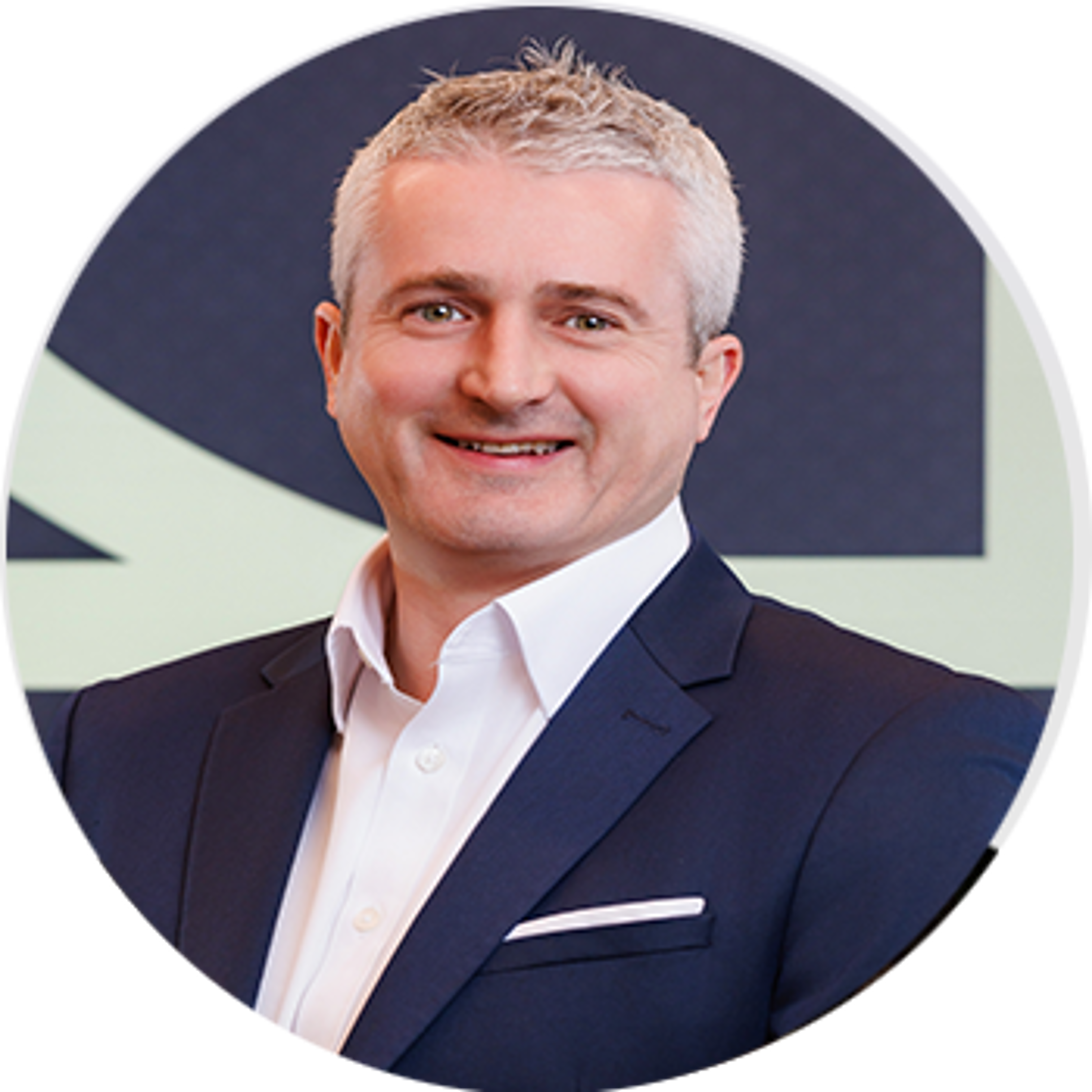A smiling man with short gray hair wearing a dark blazer, white shirt, and pocket square, posing in front of a blurred background with geometric shapes.