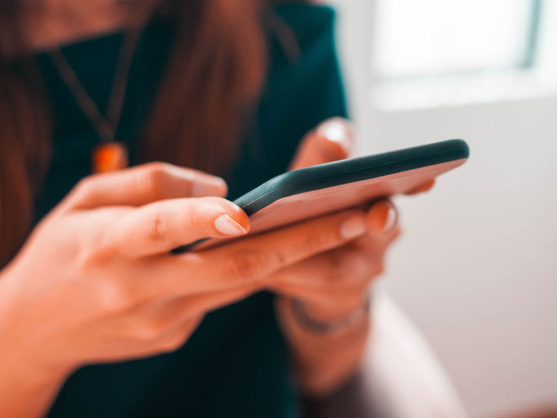 A person with long hair holds and uses a smartphone, focusing on the hands and device. The background is softly blurred, suggesting an indoor setting.