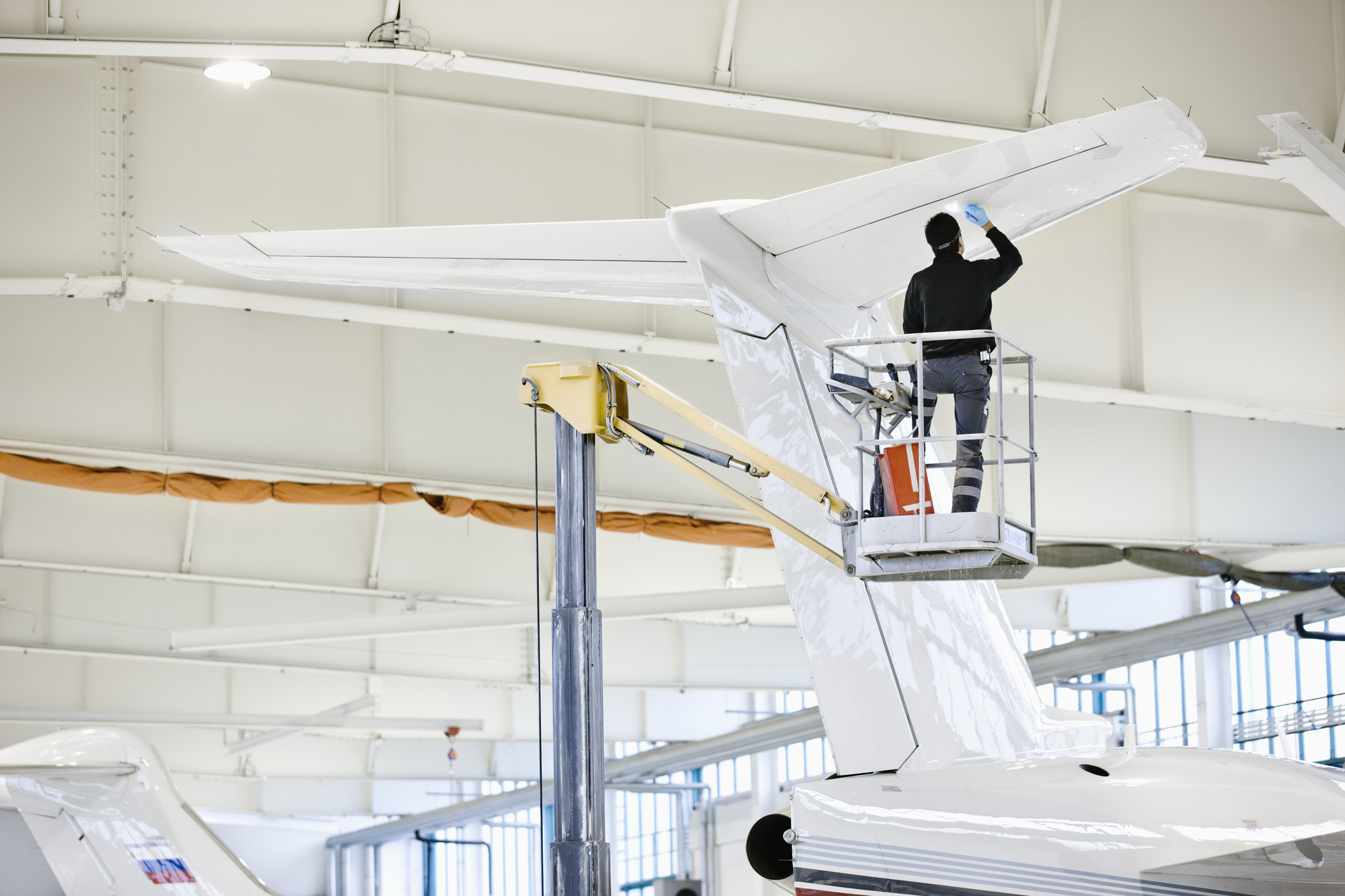 A person standing on a lift cleans or inspects the tail of a white airplane inside a bright, spacious aircraft hangar.