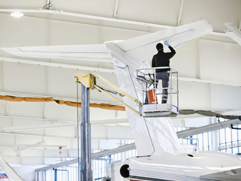 A person standing on a lift cleans or inspects the tail of a white airplane inside a bright, spacious aircraft hangar.