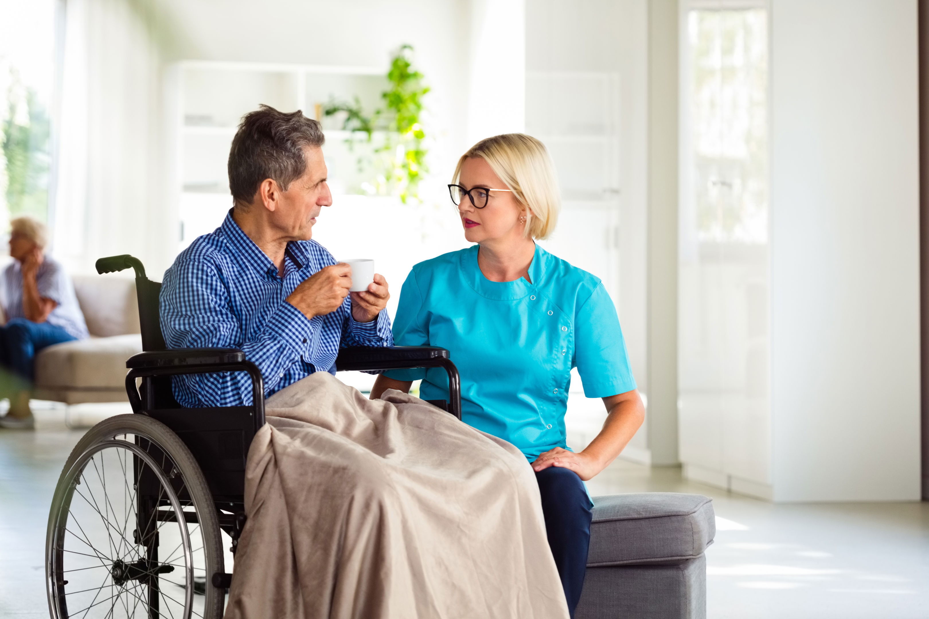 A man in a wheelchair with a blanket on his lap holds a cup and talks with a nurse in blue scrubs, who sits beside him in a bright, modern room. Another person is visible in the background.