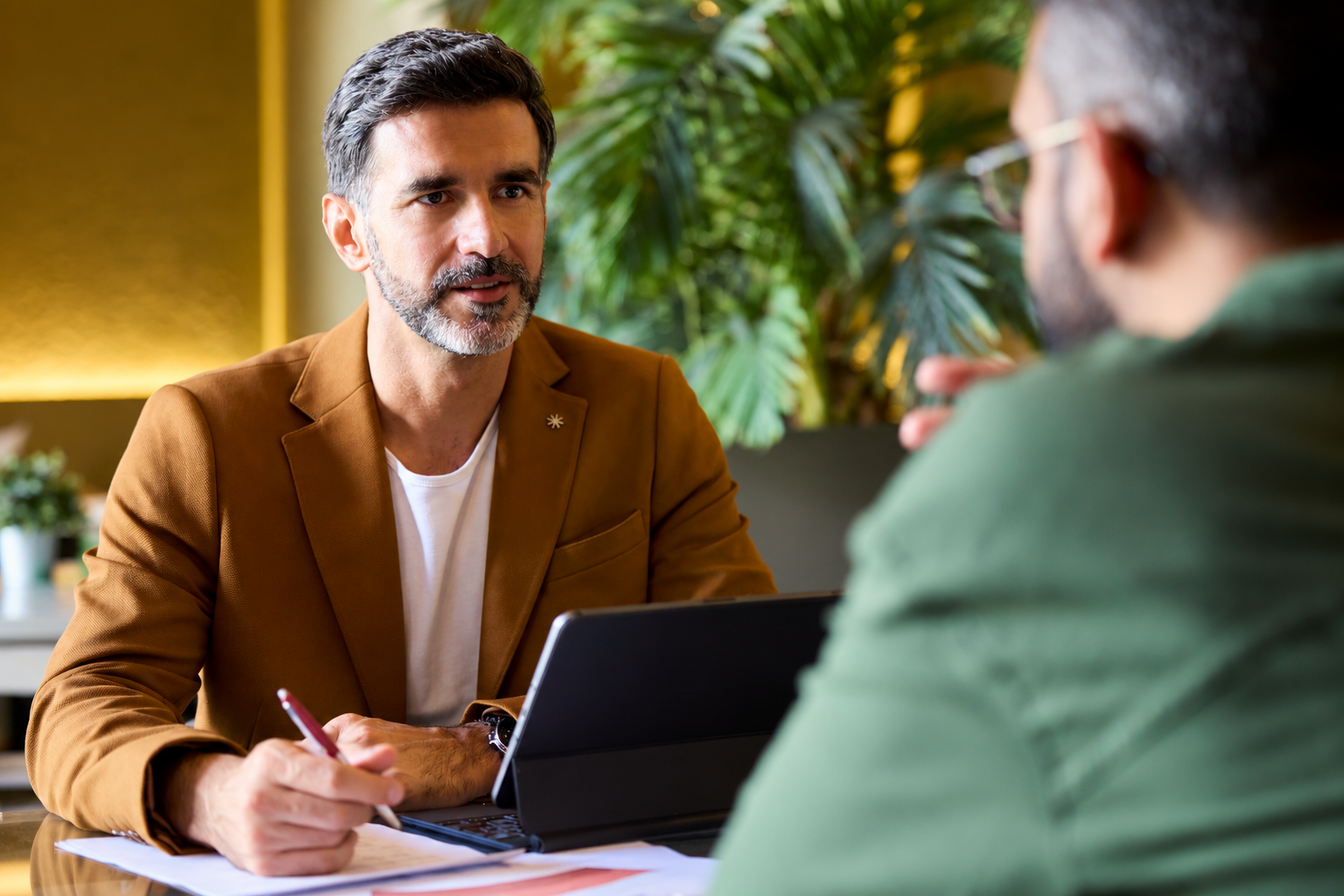 Two men sit across from each other at a desk with a laptop and documents. One man in a brown blazer listens attentively, holding a pencil, while the other man in green speaks. Green plants are in the background.