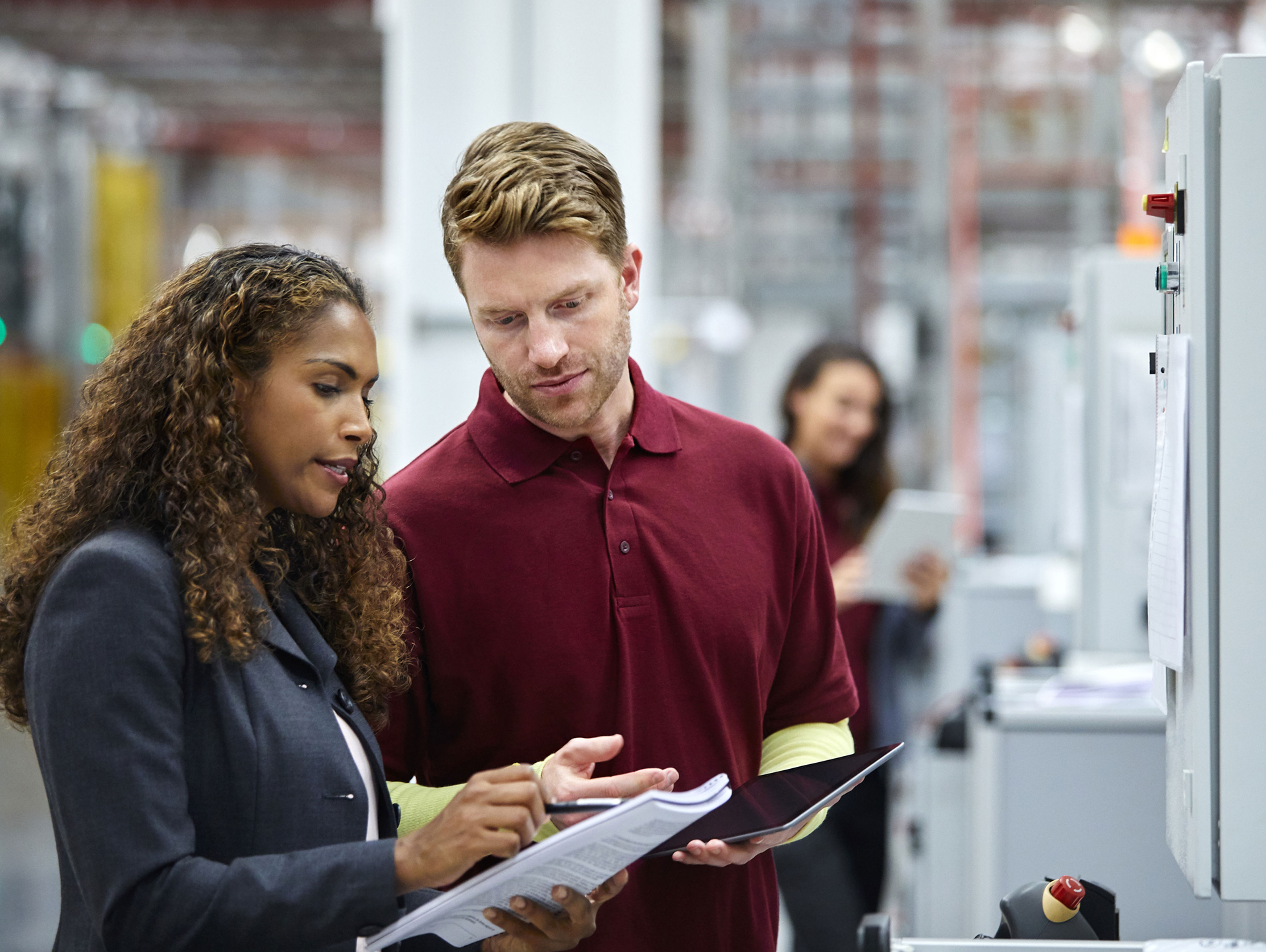 Two people in business attire discuss documents in a modern factory or industrial setting, while another person works in the background with a tablet. Machinery and equipment are visible throughout the space.
