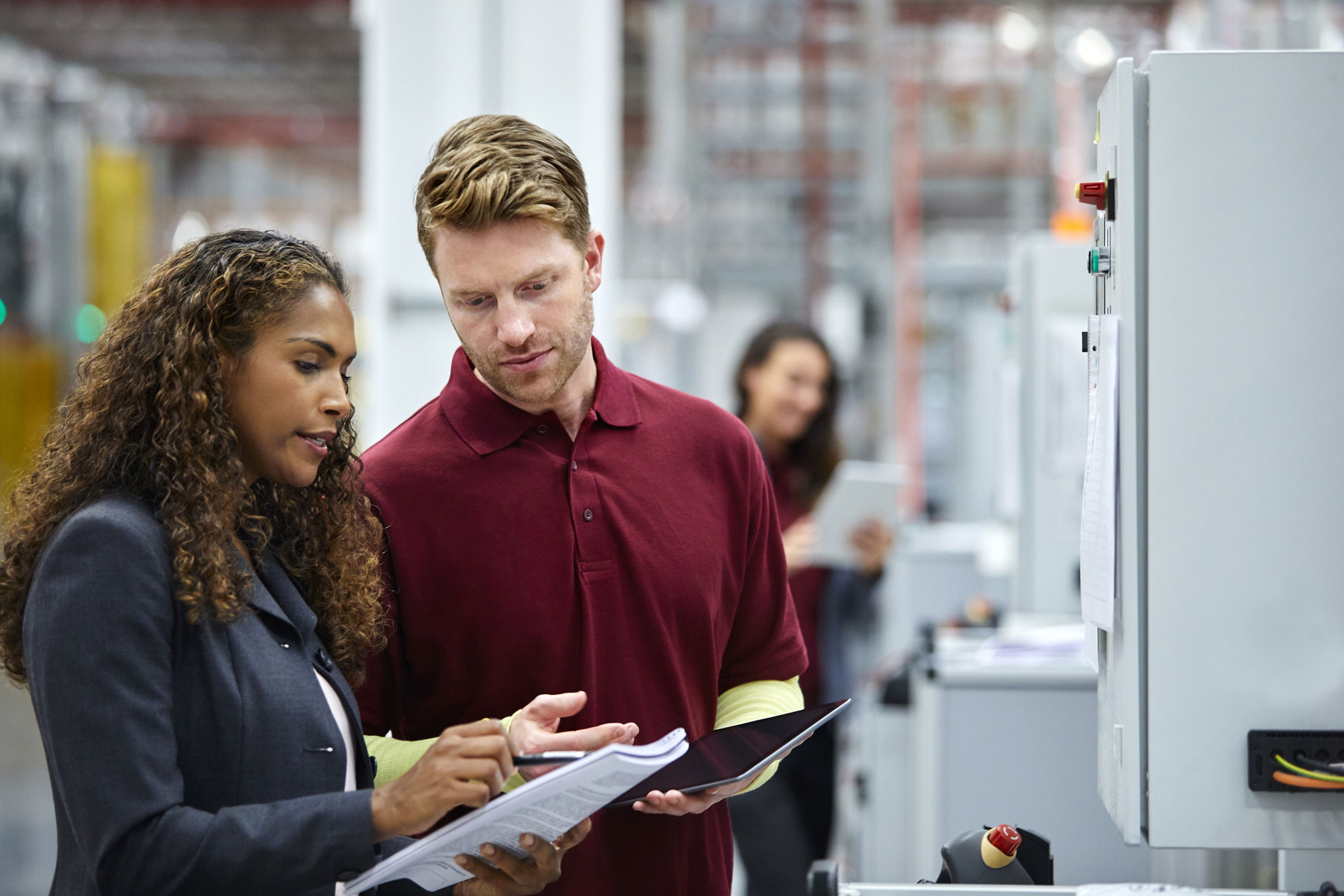 Two people in business attire discuss documents in a modern factory or industrial setting, while another person works in the background with a tablet. Machinery and equipment are visible throughout the space.