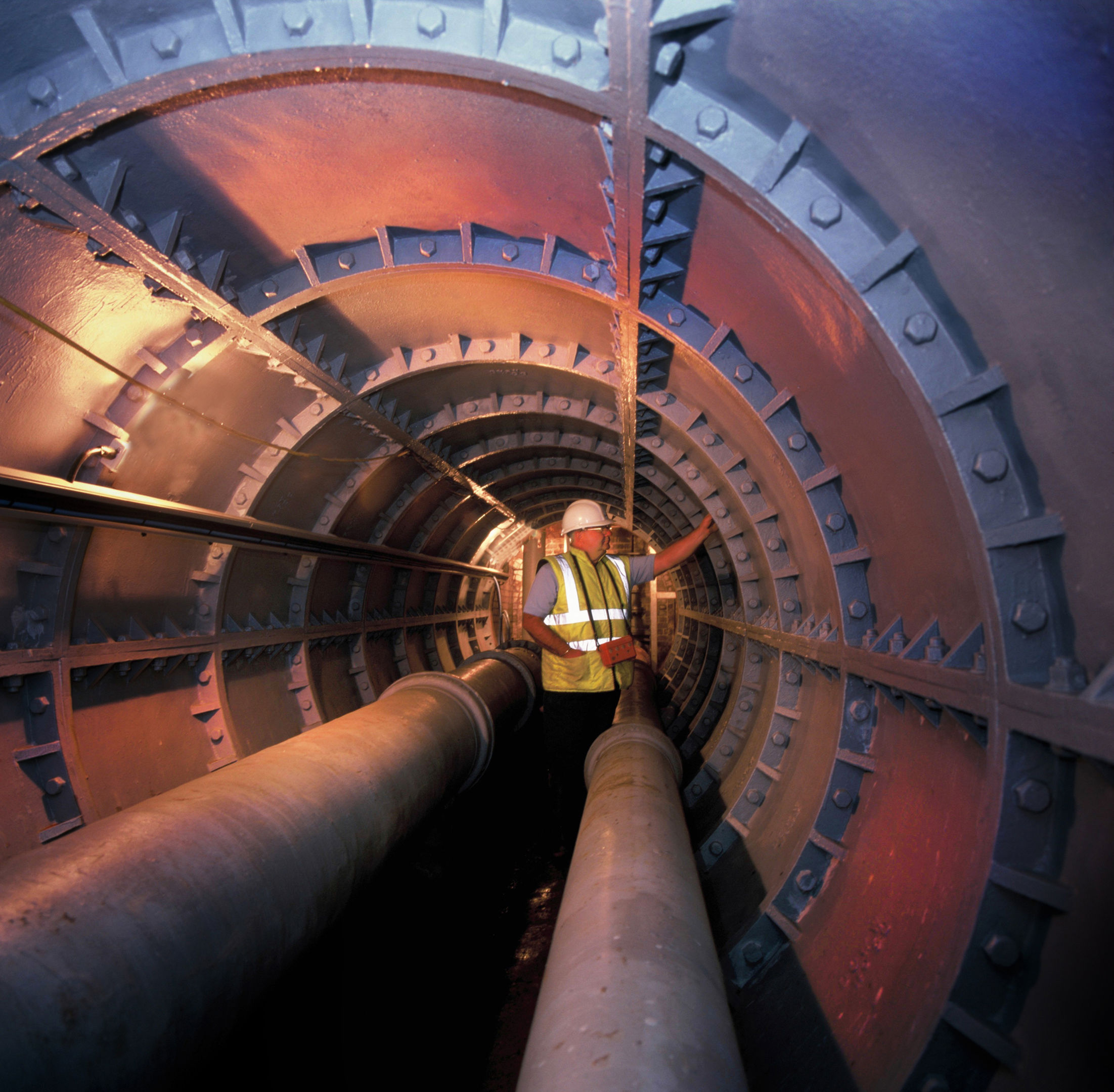 A worker wearing a hard hat and high-visibility vest stands inside a large, round industrial tunnel lined with metal panels and pipes, illuminated by artificial lighting.