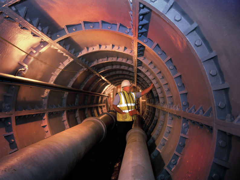 A worker wearing a hard hat and high-visibility vest stands inside a large, round industrial tunnel lined with metal panels and pipes, illuminated by artificial lighting.