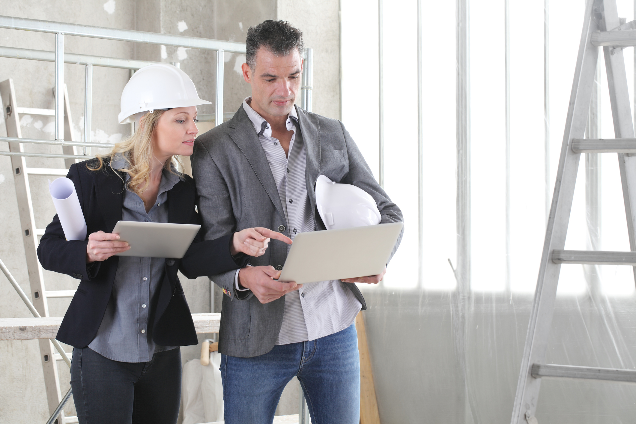 Two professionals, one wearing a hard hat, stand in a construction site. They are looking at a laptop and a tablet, discussing plans. Construction materials and a ladder are visible in the background.