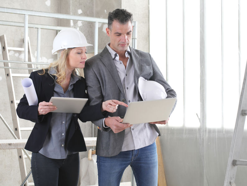 Two professionals, one wearing a hard hat, stand in a construction site. They are looking at a laptop and a tablet, discussing plans. Construction materials and a ladder are visible in the background.