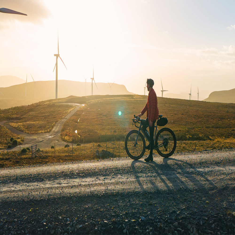 A person stands with a bicycle on a gravel road at sunset, overlooking a landscape with wind turbines and rolling hills under a bright sky.