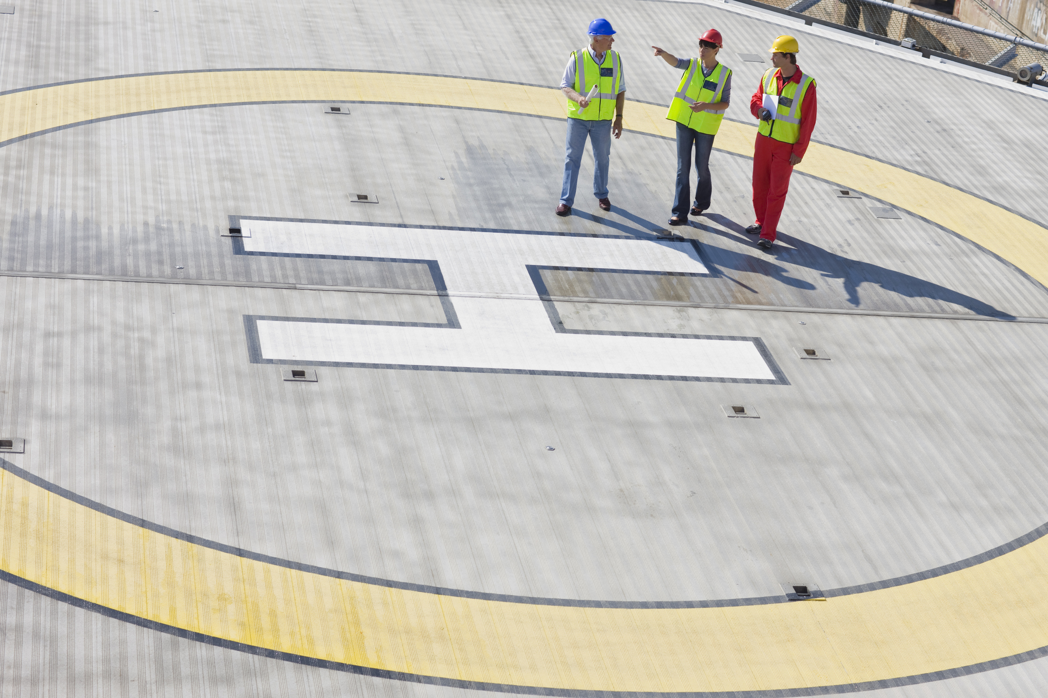 Three construction workers in safety gear and helmets stand on a large helipad marked with a white “H” inside a yellow circle, talking and reviewing documents.