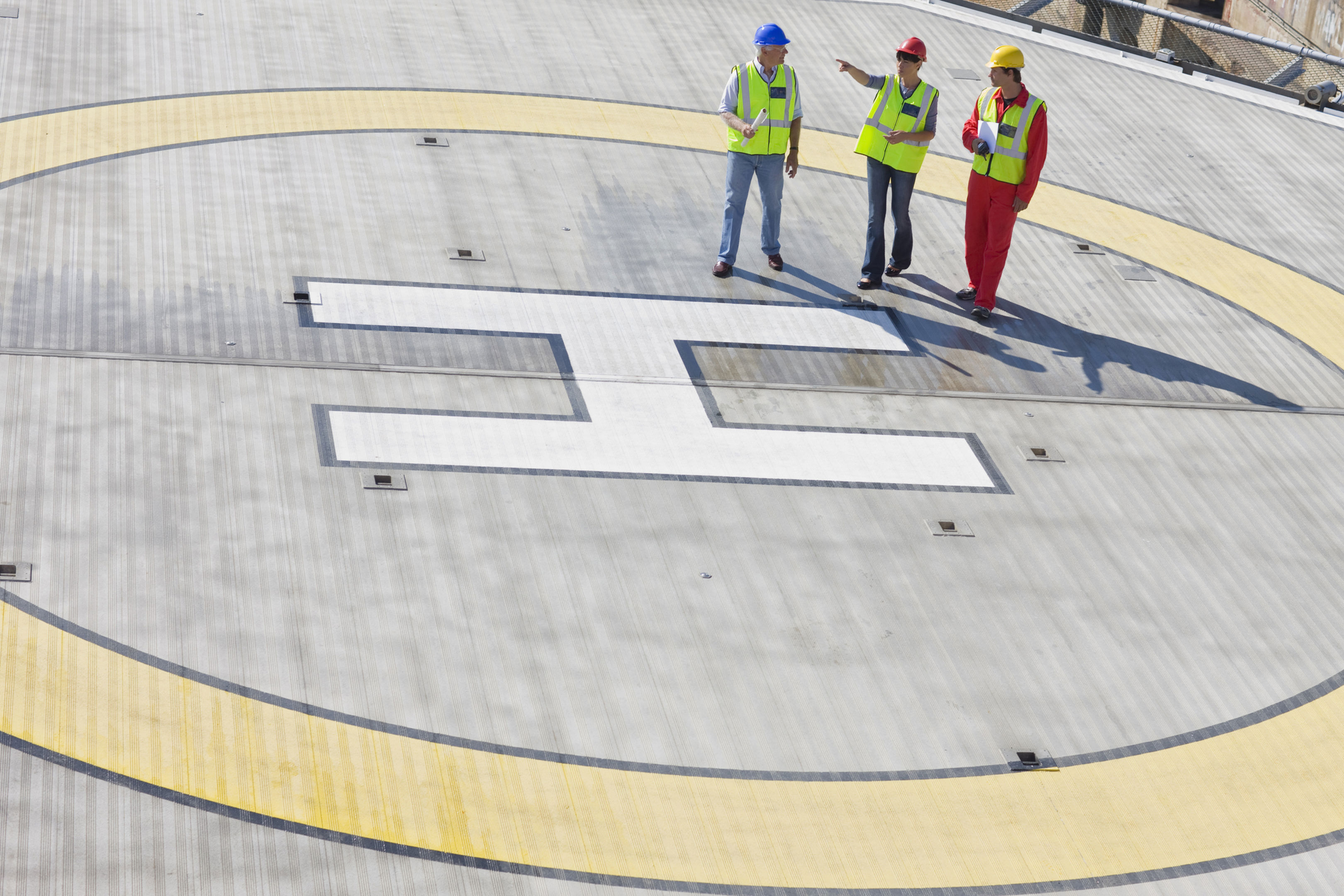 Three construction workers in safety gear and helmets stand on a large helipad marked with a white “H” inside a yellow circle, talking and reviewing documents.