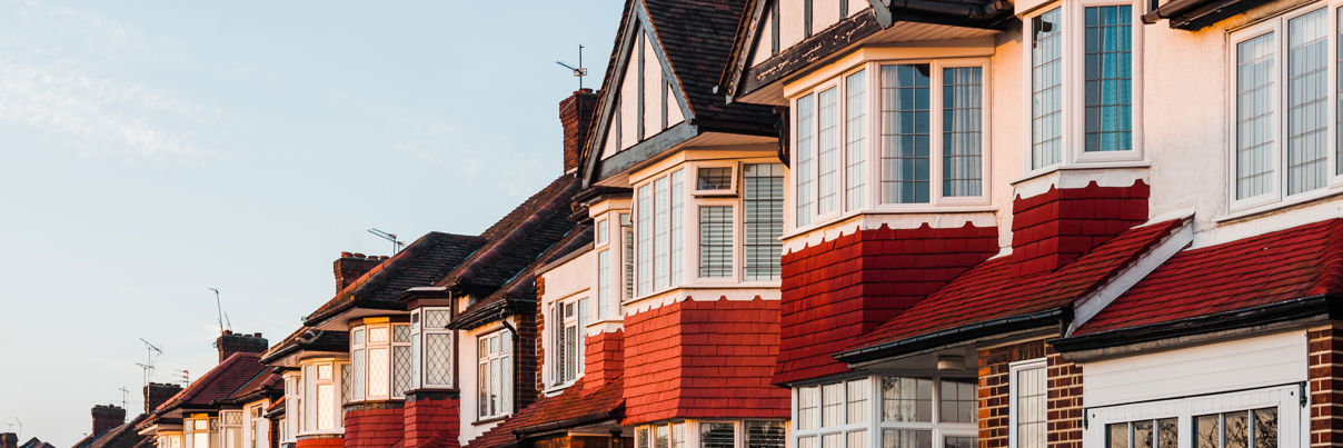 A row of traditional English-style houses with red brick and white accents. The homes have large bay windows, steep gabled roofs, and small chimneys. The sky is clear and blue, suggesting a sunny day.