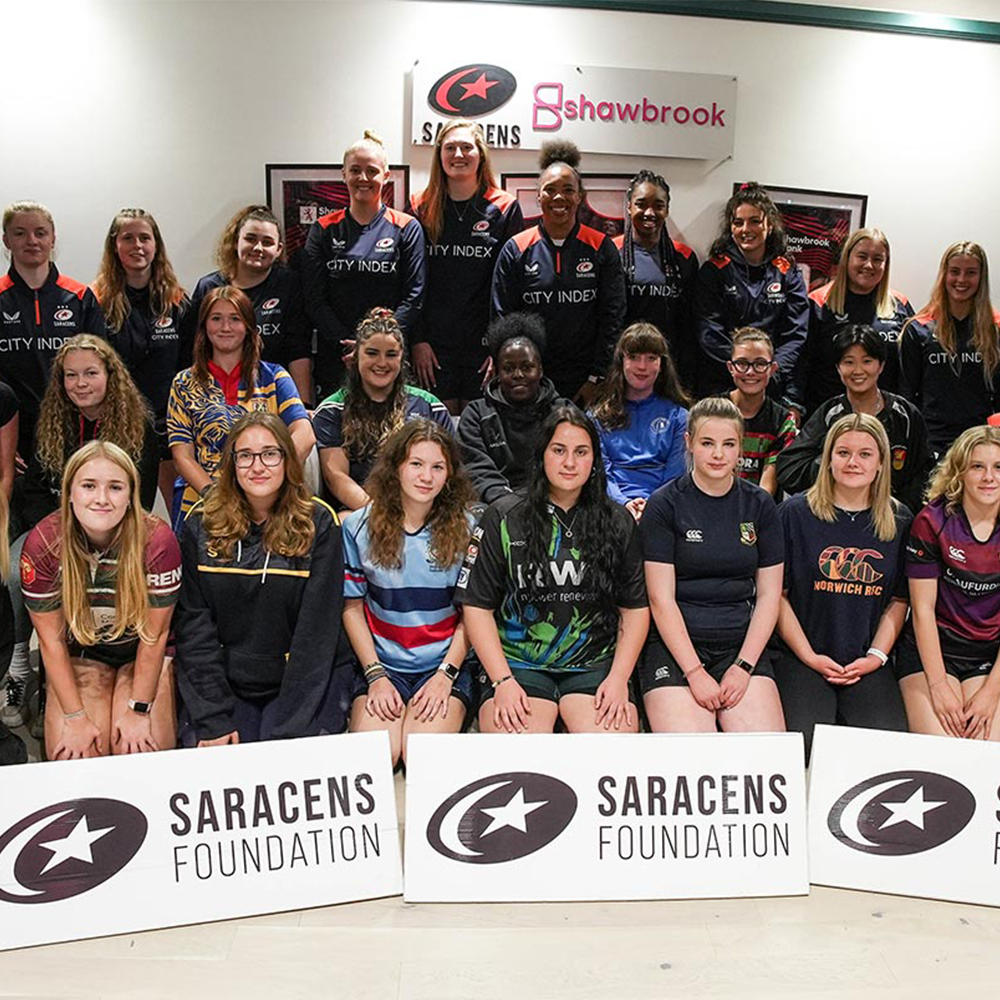 A group of women and girls pose indoors wearing various rugby jerseys, standing and sitting behind Saracens Foundation signs, with a Saracens banner on the wall behind them.