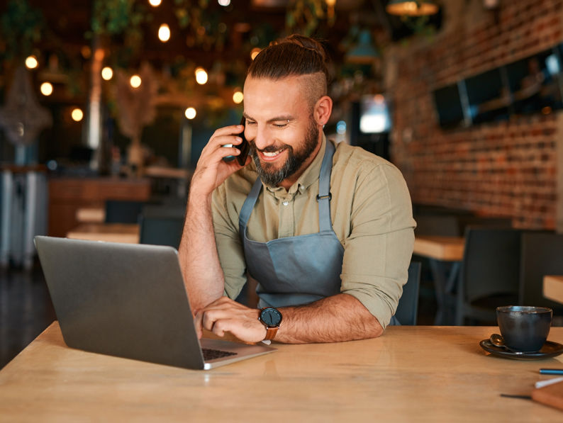 A smiling man in an apron sits at a table in a cafe, using a laptop and talking on his phone. A cup of coffee is on the table beside him. The cafe has warm lighting and a cozy atmosphere.
