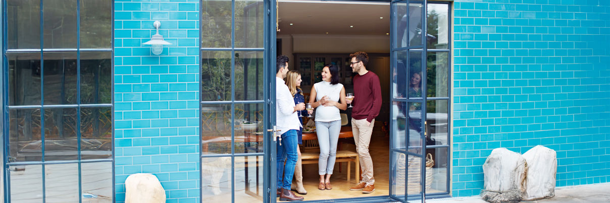 A person with long hair, wearing a jacket and backpack, stands with arms raised facing a lake and mountains under a blue sky with clouds, suggesting a sense of freedom and adventure.