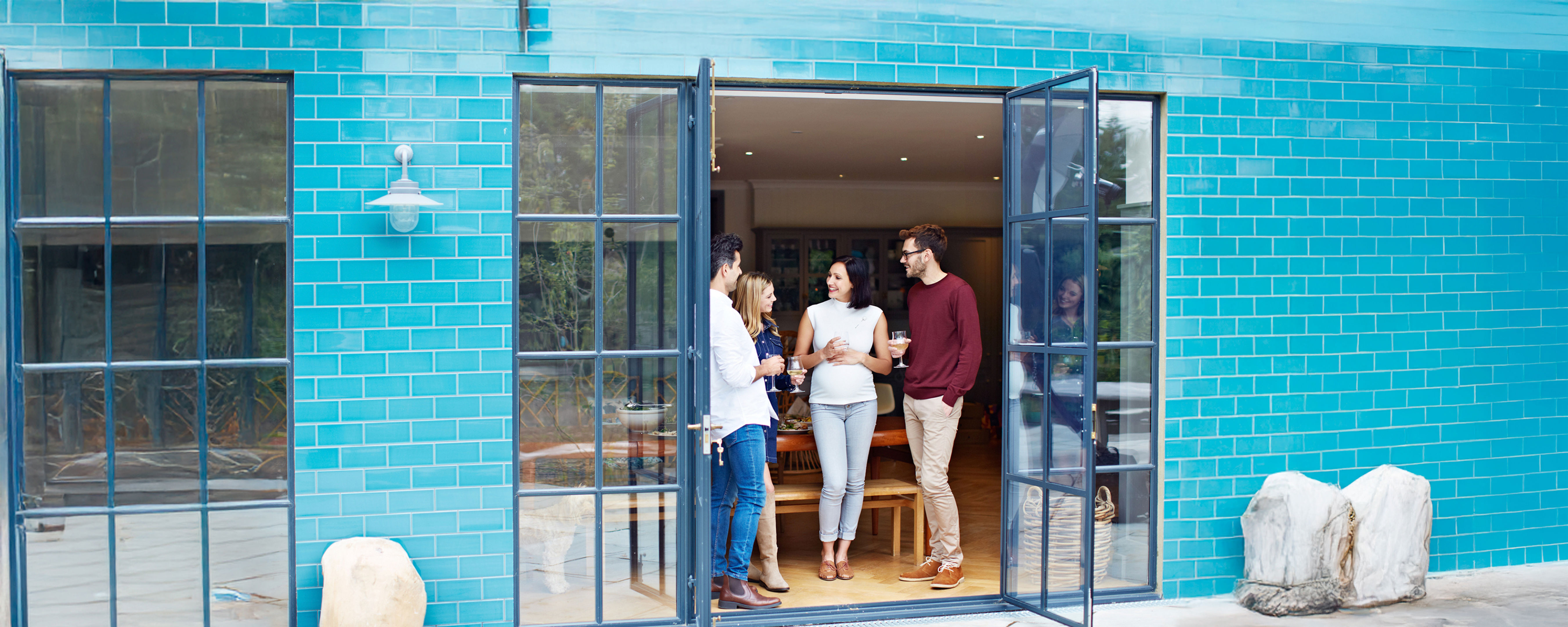 A person with long hair, wearing a jacket and backpack, stands with arms raised facing a lake and mountains under a blue sky with clouds, suggesting a sense of freedom and adventure.