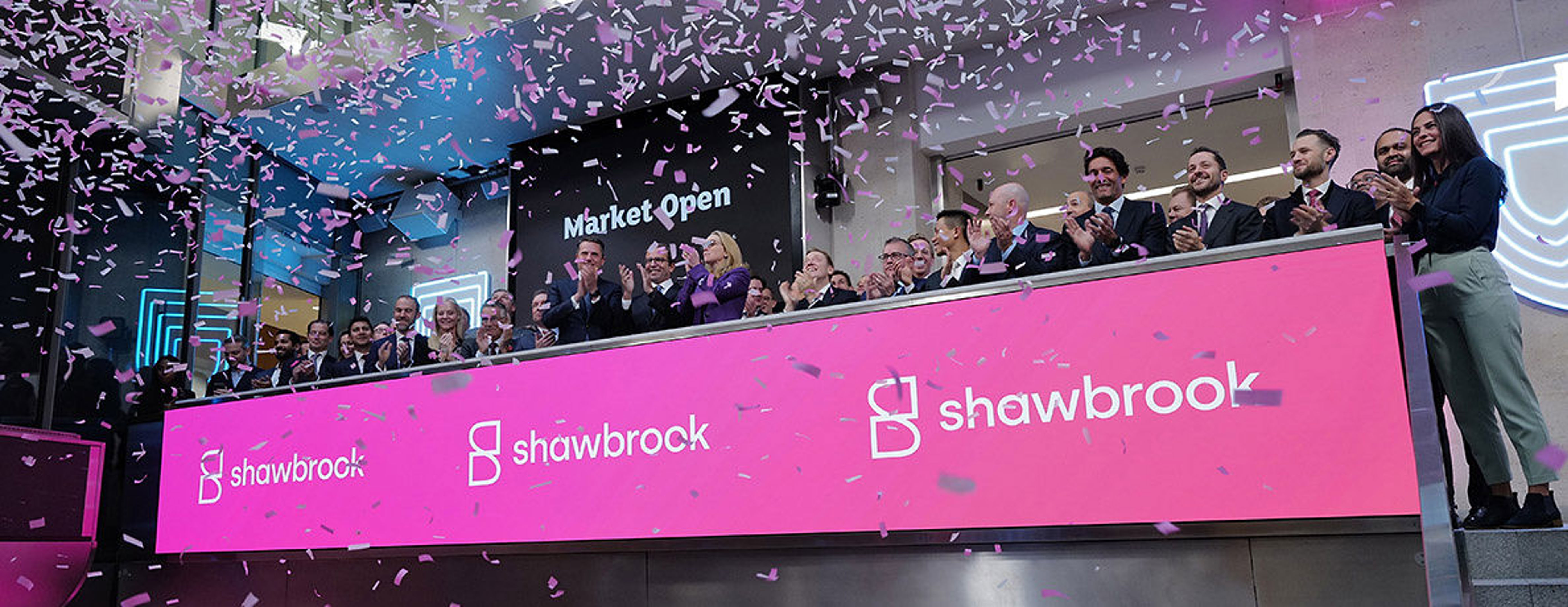 A group of people stand behind a large pink sign with the Shawbrook logo, clapping and celebrating as pink confetti falls, with "Market Open" displayed in the background.