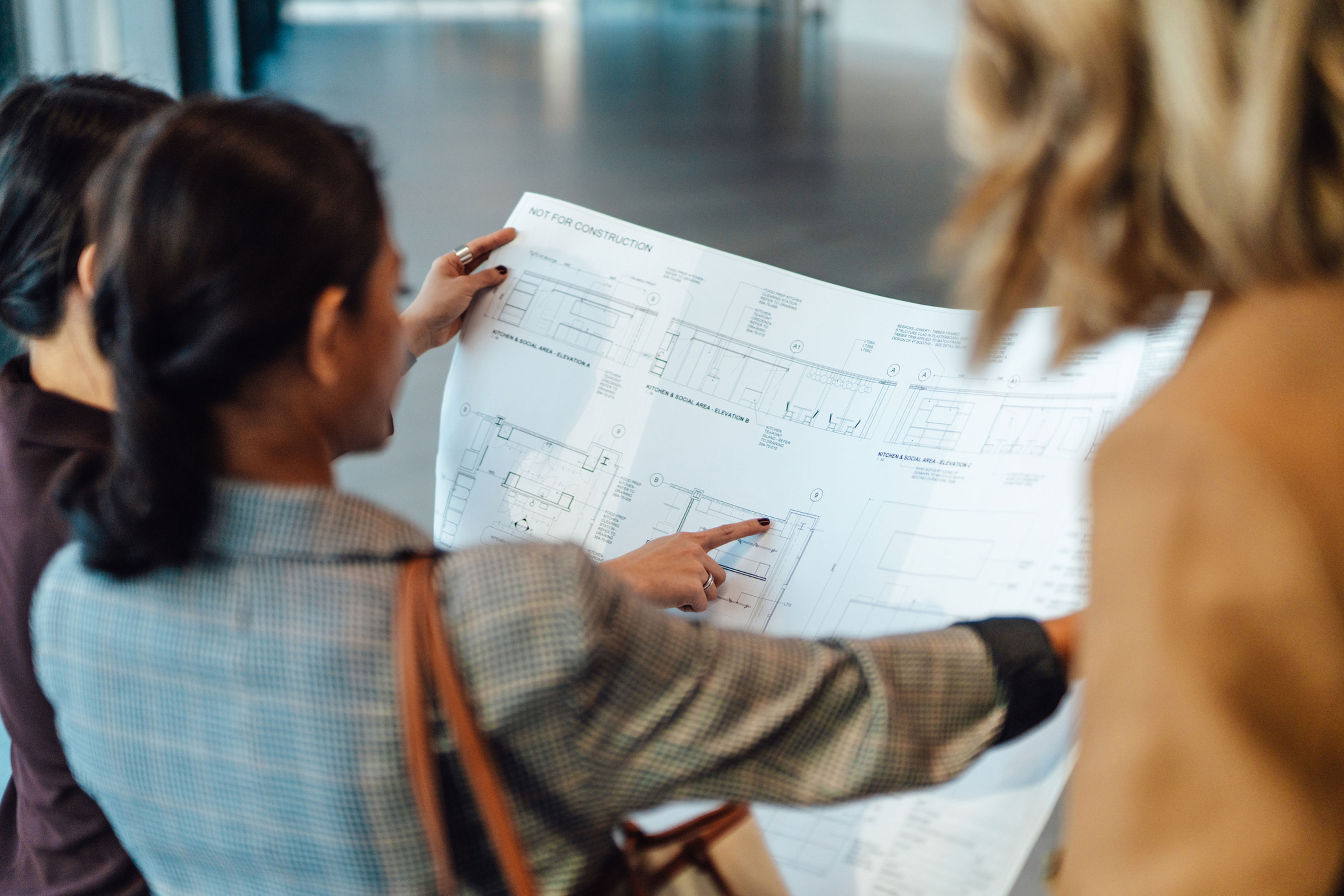Three women stand together, closely examining and pointing at architectural blueprints or construction plans in a well-lit indoor space.