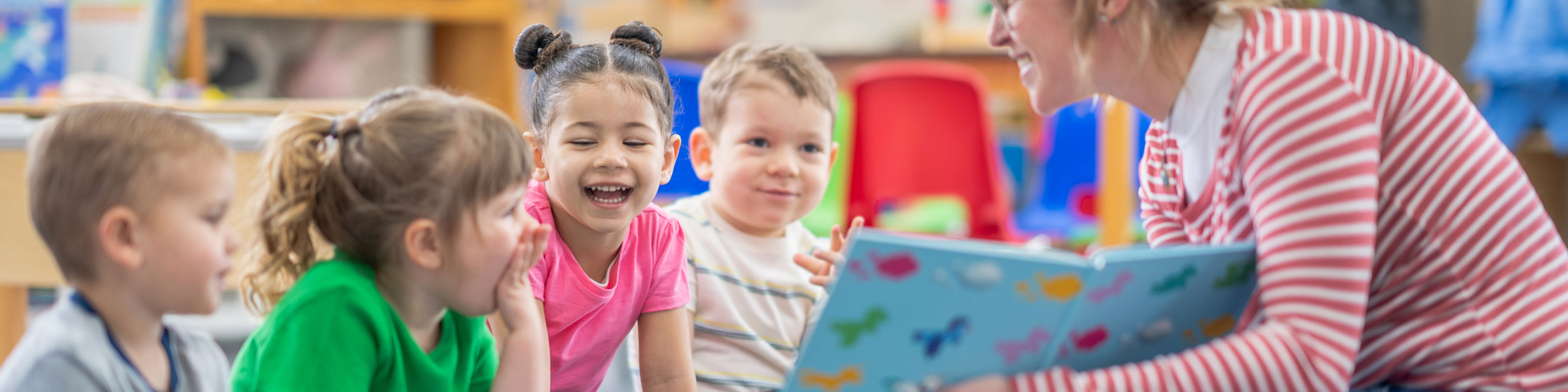 A woman reads a colorful book to a group of four young children sitting on the floor in a cheerful classroom, all smiling and engaged.