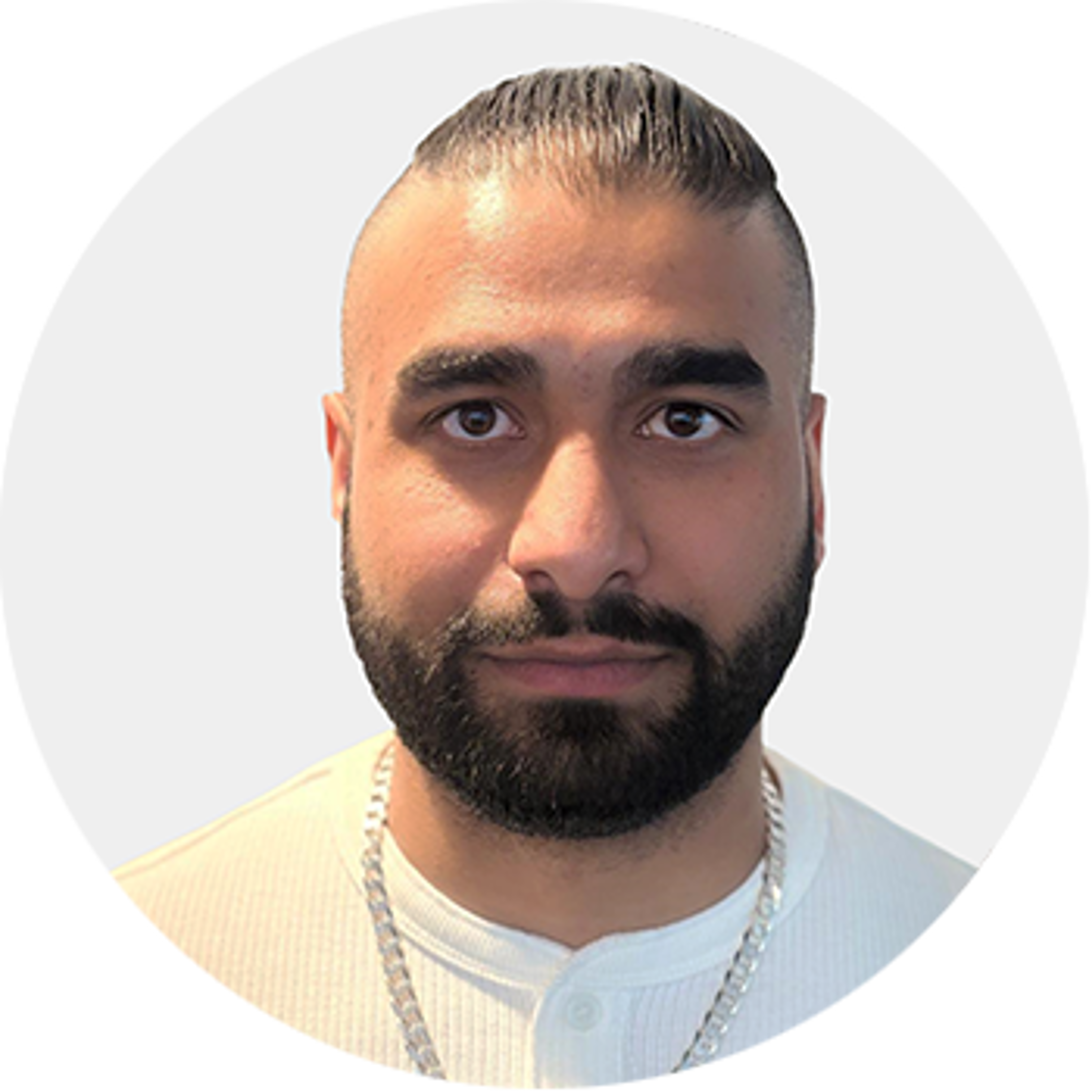 A man with a beard and mustache, wearing a white shirt and a silver chain necklace, stands against a light gray background. His hair is styled and slicked back on top.