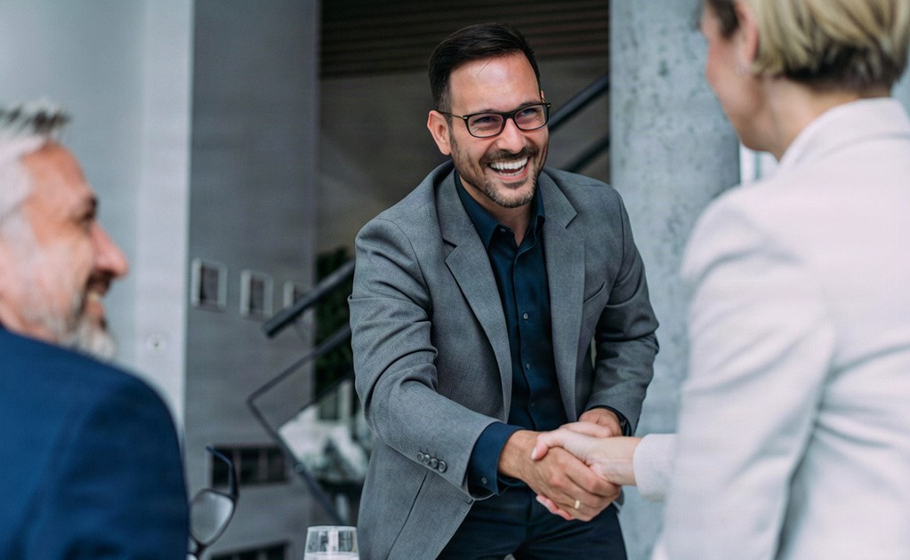 A man in a grey blazer smiling and shaking hands with a woman in a professional setting.