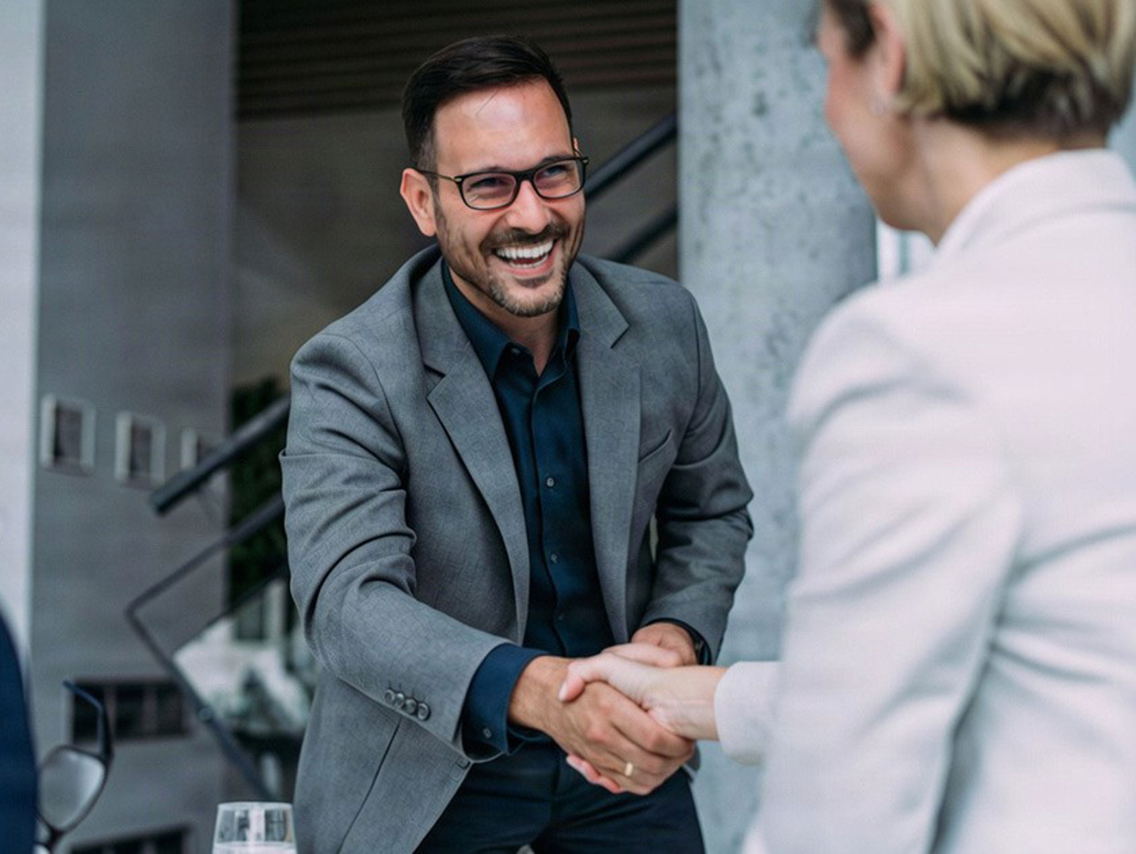 A man in a grey blazer smiling and shaking hands with a woman in a professional setting.