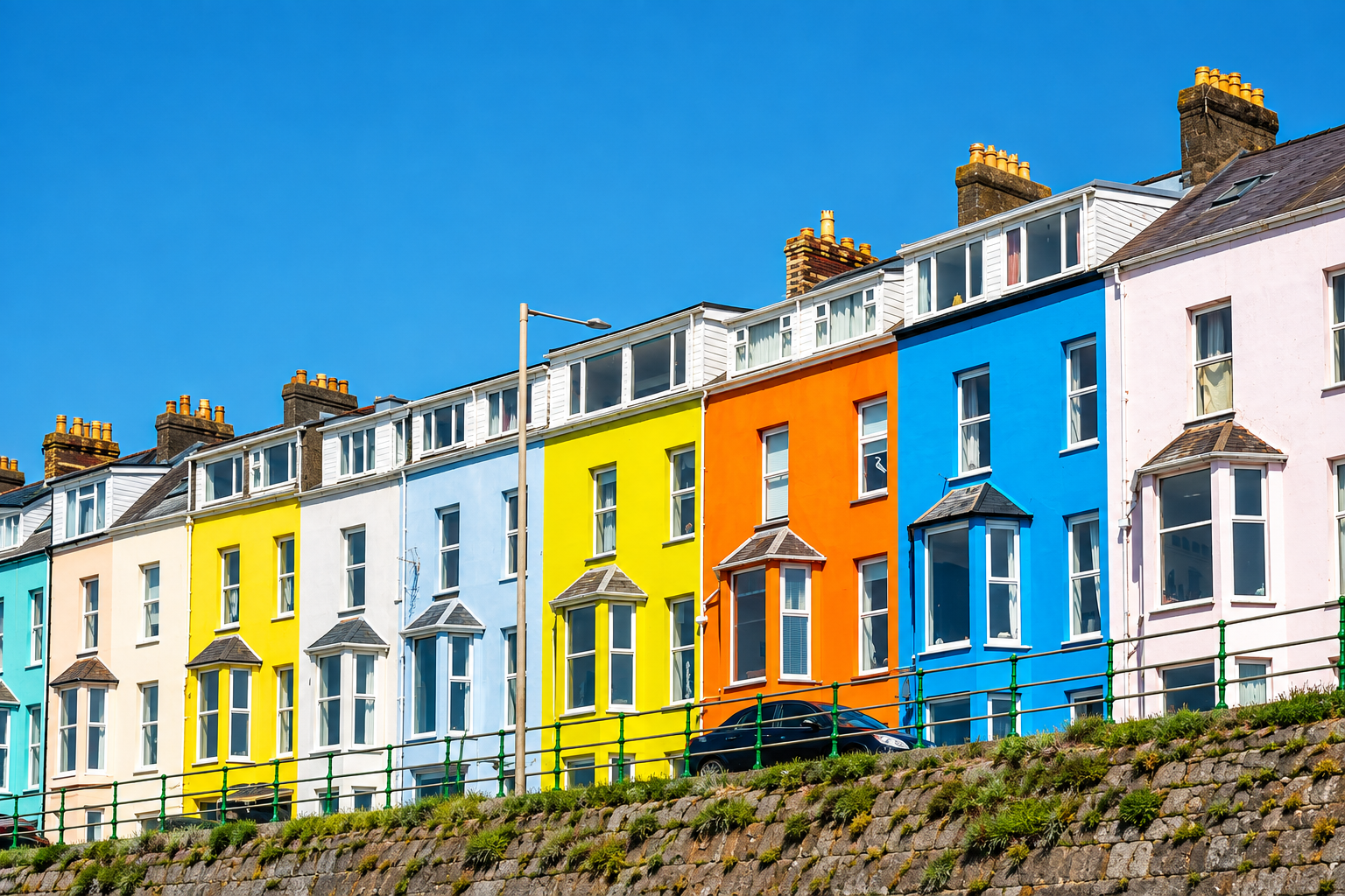 A row of colorful, multi-story terraced houses with bay windows under a clear blue sky, seen behind a stone wall and green railings.