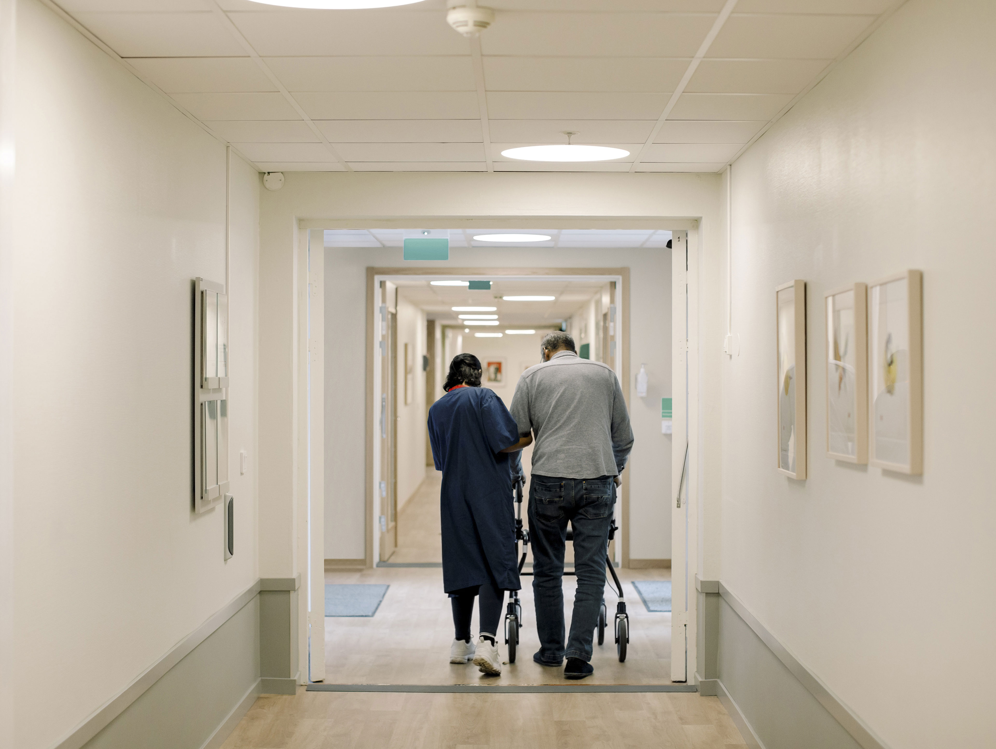 Nurse Walking With Senior Man In Corridor At Nursing Home