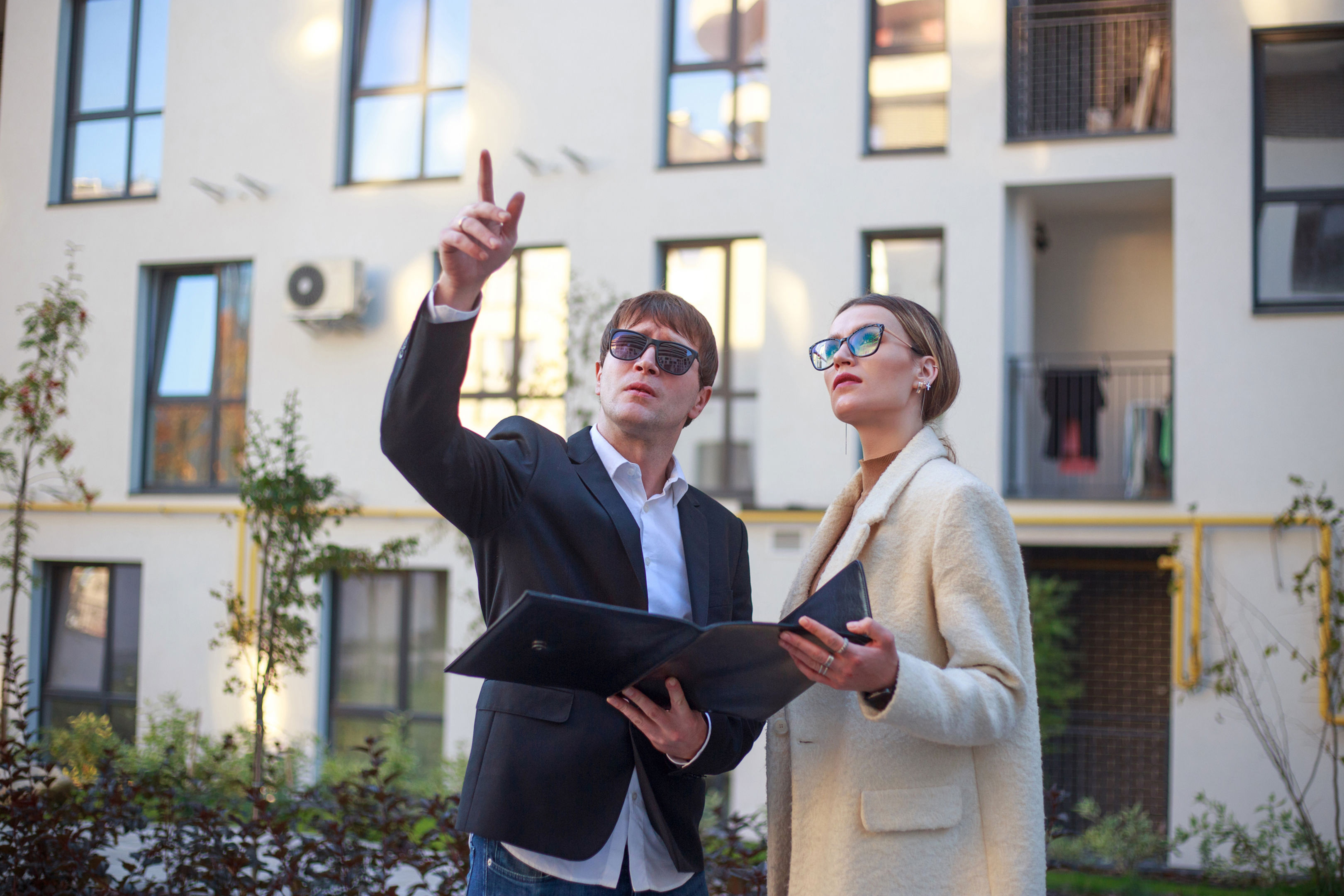 A man and woman in business attire stand outside a modern apartment building. The man, wearing sunglasses and a blazer, points upward, while the woman, also in sunglasses and a coat, holds an open folder, looking in the same direction.